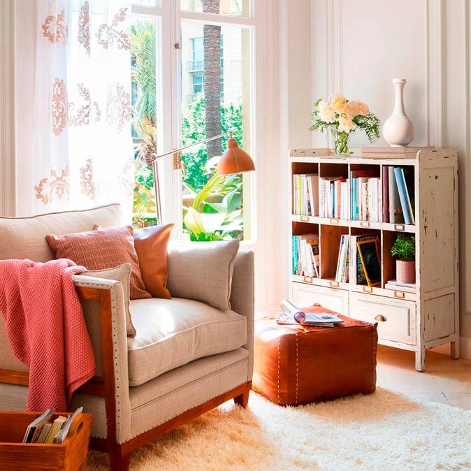 Butaca beige con estructura de madera, librería pequeña, puff de piel, lámpara de pie tipo flexo y alfombra de pelo en crudo