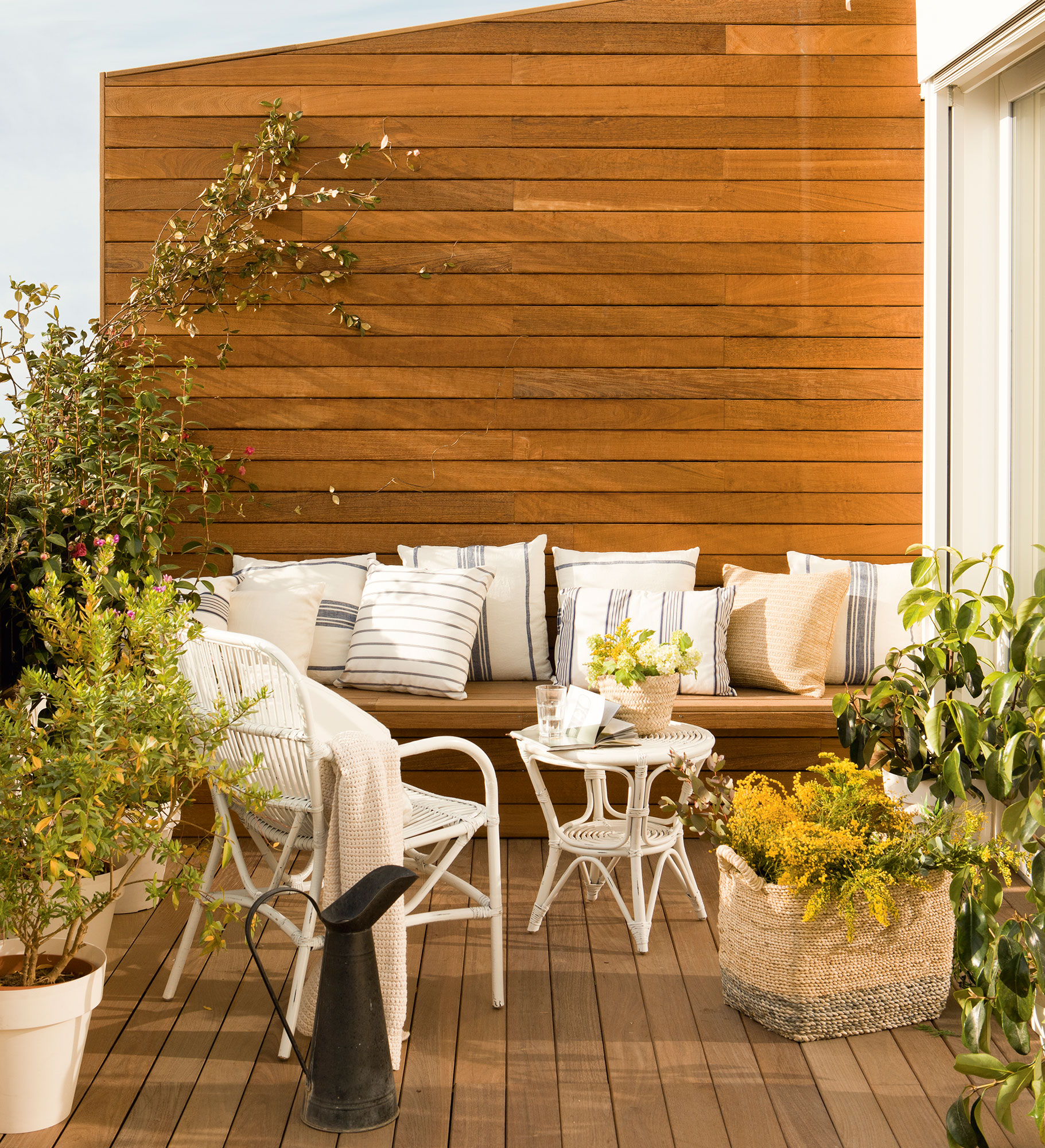 Terraza con banco de madera integrado en la pared y decorado con cojines frente a una mesa y una butaca de fibras en color blanco