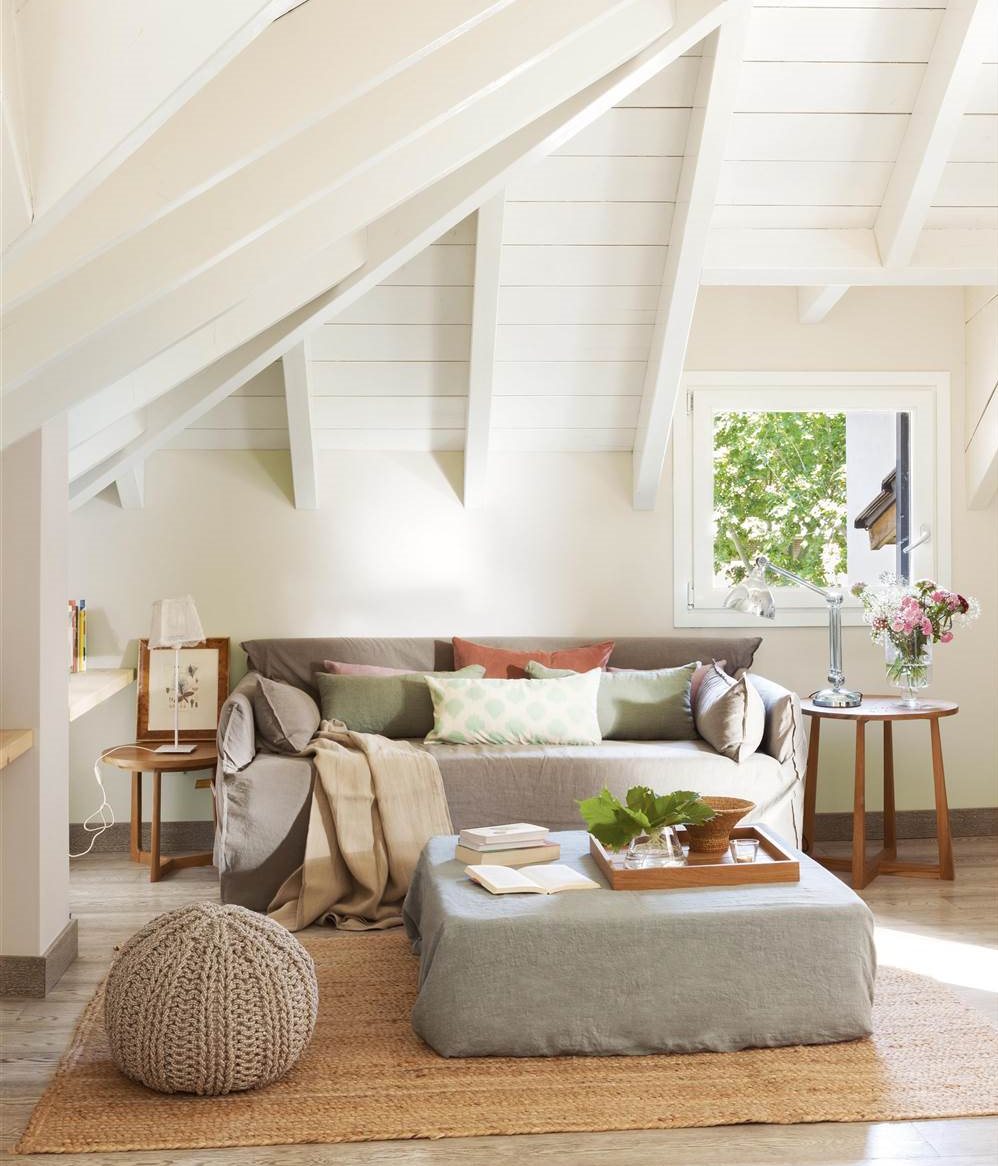 Salón en espacio con techo abuhardillado blanco, sofá y reposapiés-mesa de centro en gris con fundas, puf de tricot redondo, mesitas auxiliares de madera redondas y alfombra