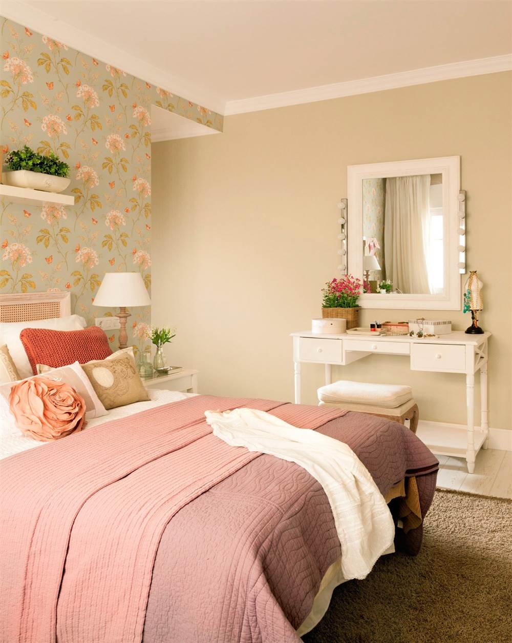 Dormitorio con papel pintado verde con flores, ropa de cama rosa viejo y muchos cojines, tocador con banqueta y espejo con luces de camerino