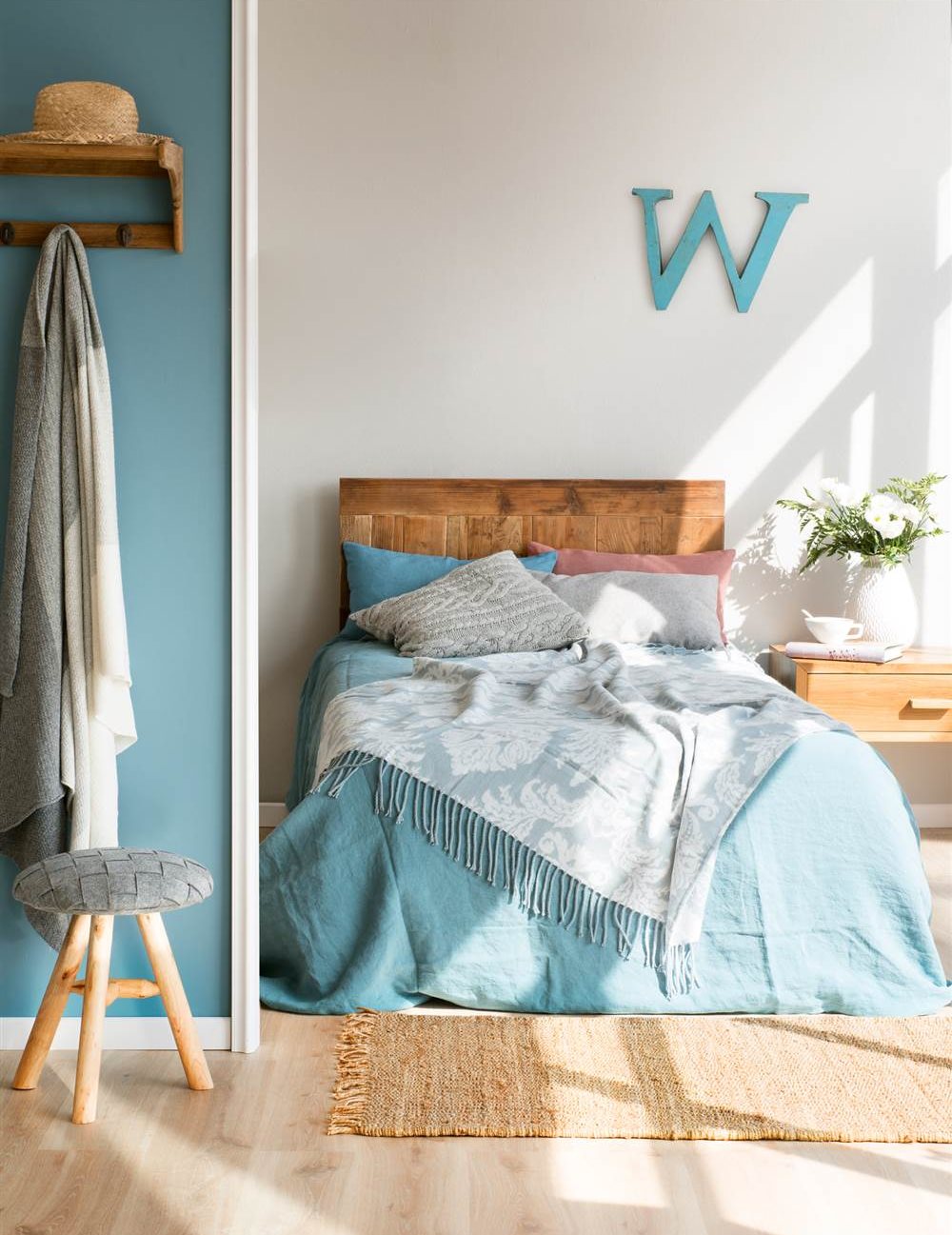 Dormitorio con pared gris piedra y azul, con cabecero y mesita de noche de madera, letra W decorativa, ropa de cama con estampado damasco, taburete y colgador