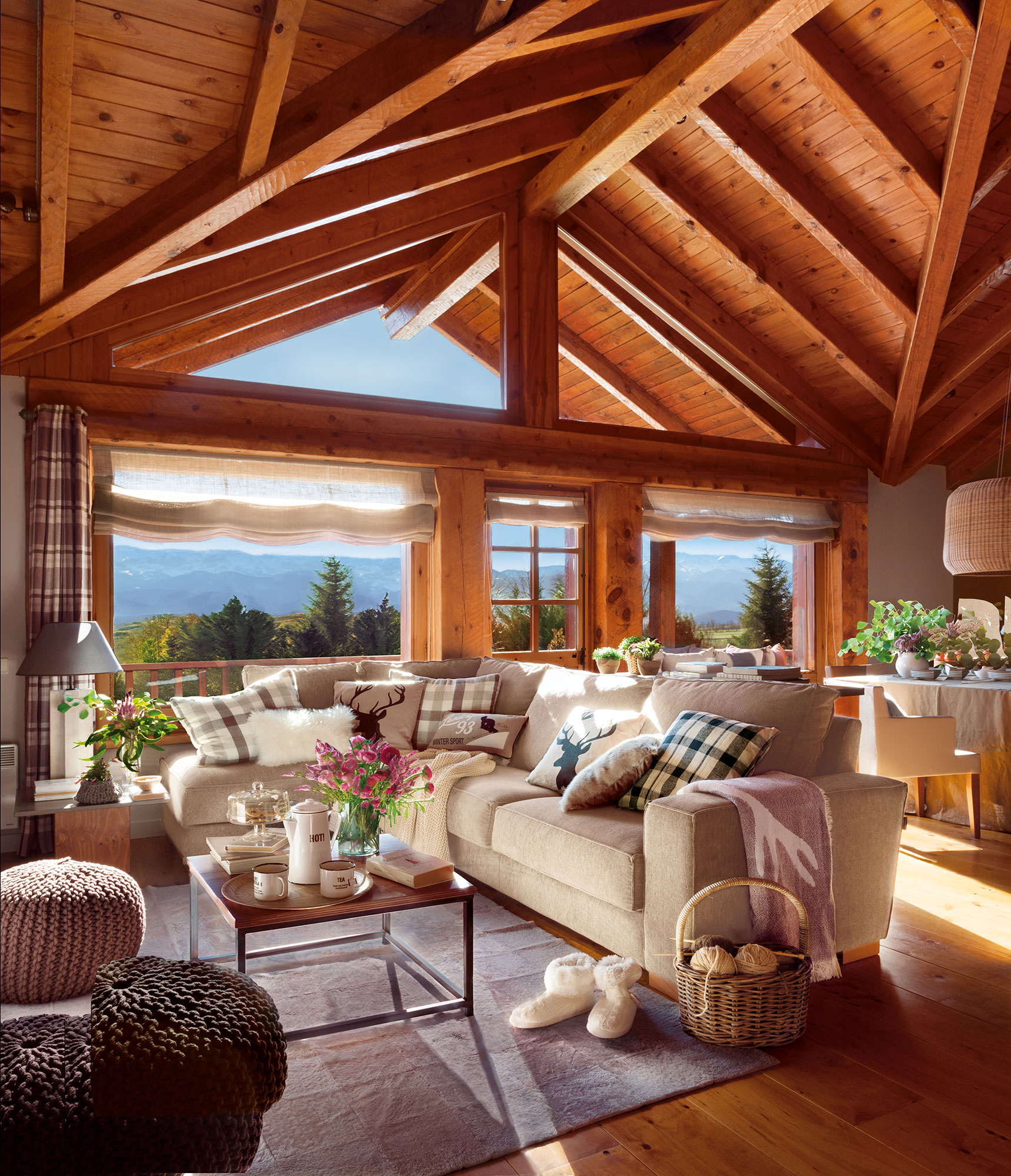 Salón con techos de madera abuhardillados, sofá rinconero beige y grandes ventanas. 