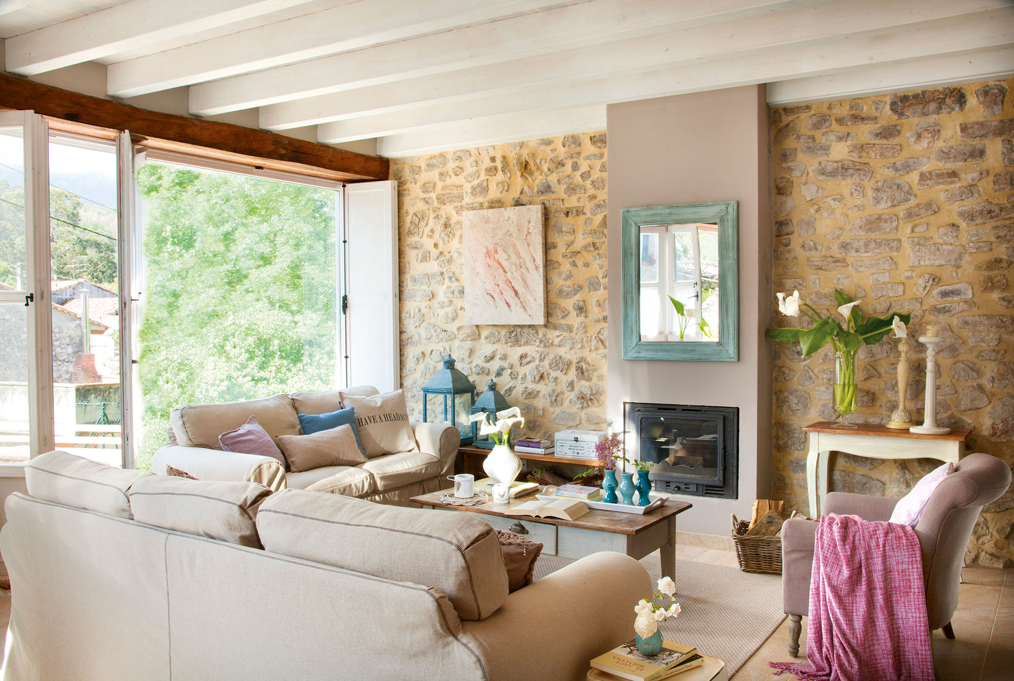 Salón con paredes de piedra, chimenea, sofás beige y gran ventanal. 