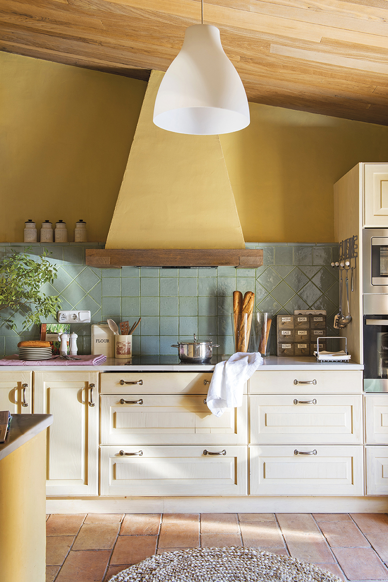 Cocina rústica con paredes amarillas, antepecho de azulejos verdes y muebles en crema. 