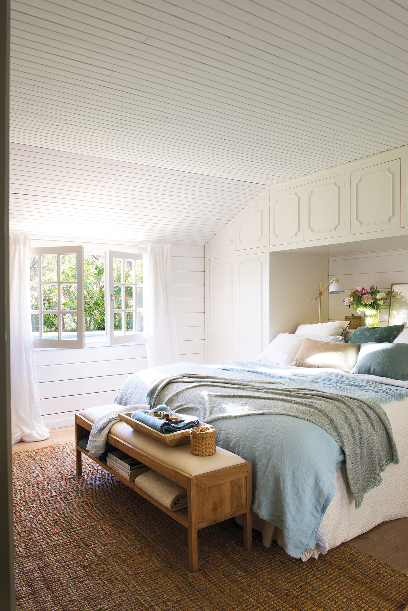 Dormitorio rústico con alfombra, banco al pie de la cama, cabecero a medida con armario y panelado de madera como revestimiento.