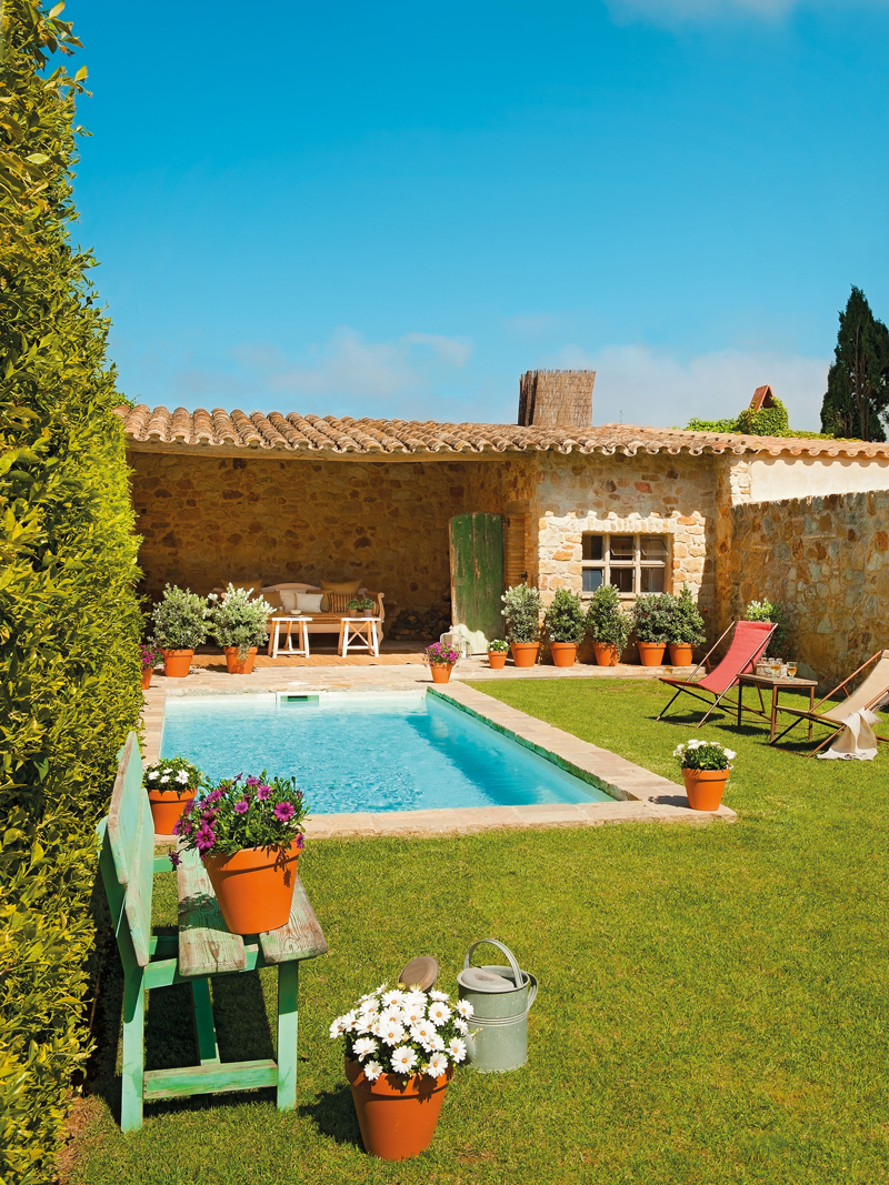 Jardín con piscina rodeada de macetas con plantas de exterior, un banquito y dos tumbonas con mesita auxiliar