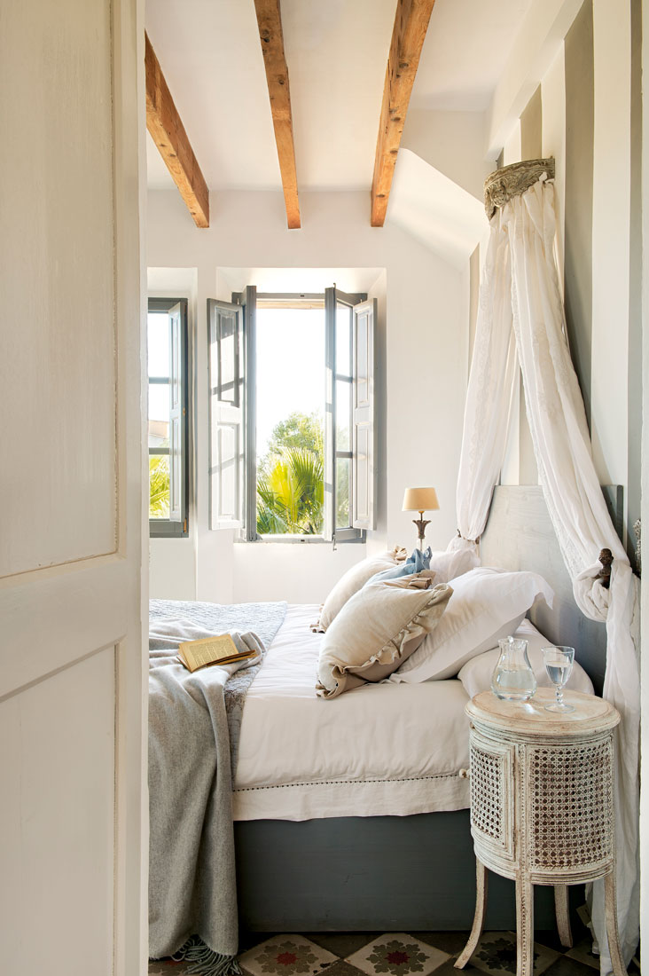 Dormitorio de estilo vintage con cama con dosel de tela blanca y mesilla de noche antigua con pintura decapada.