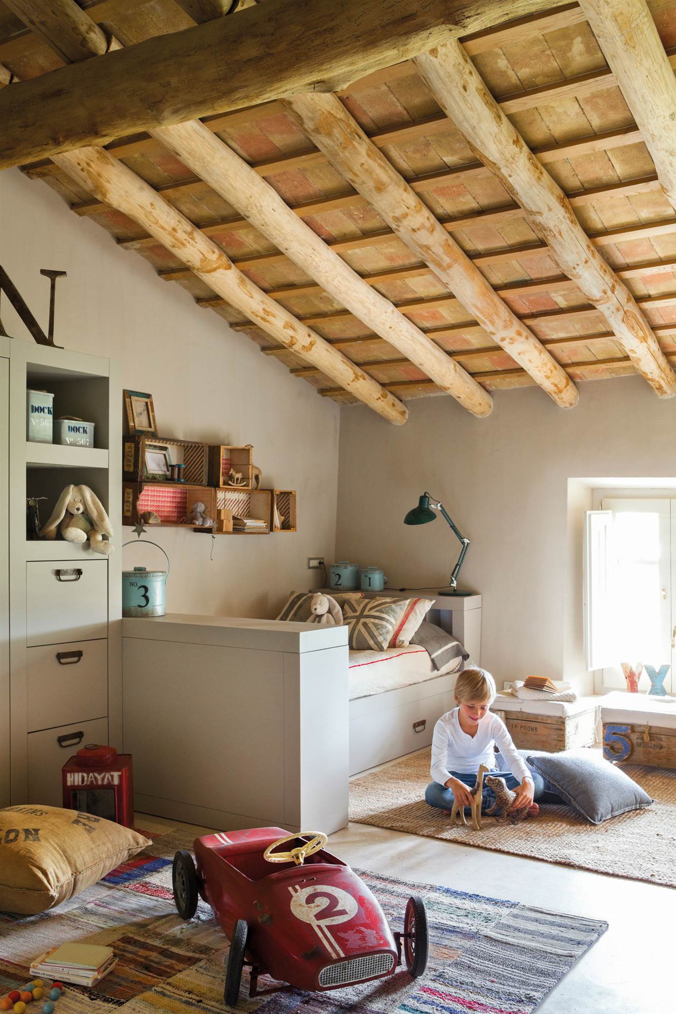 Dormitorio infantil abuhardillado con vigas de madera, alfombras, murete gris y coche de juguete.