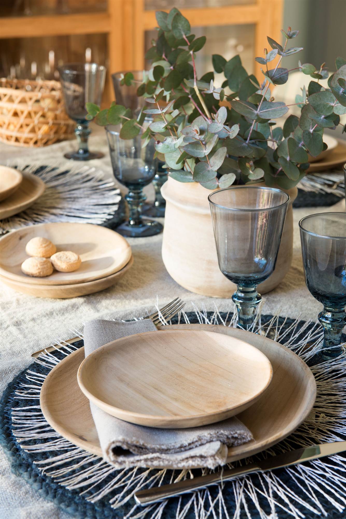 Mesa con vajilla de madera y copas azules sobre mantel de lino.