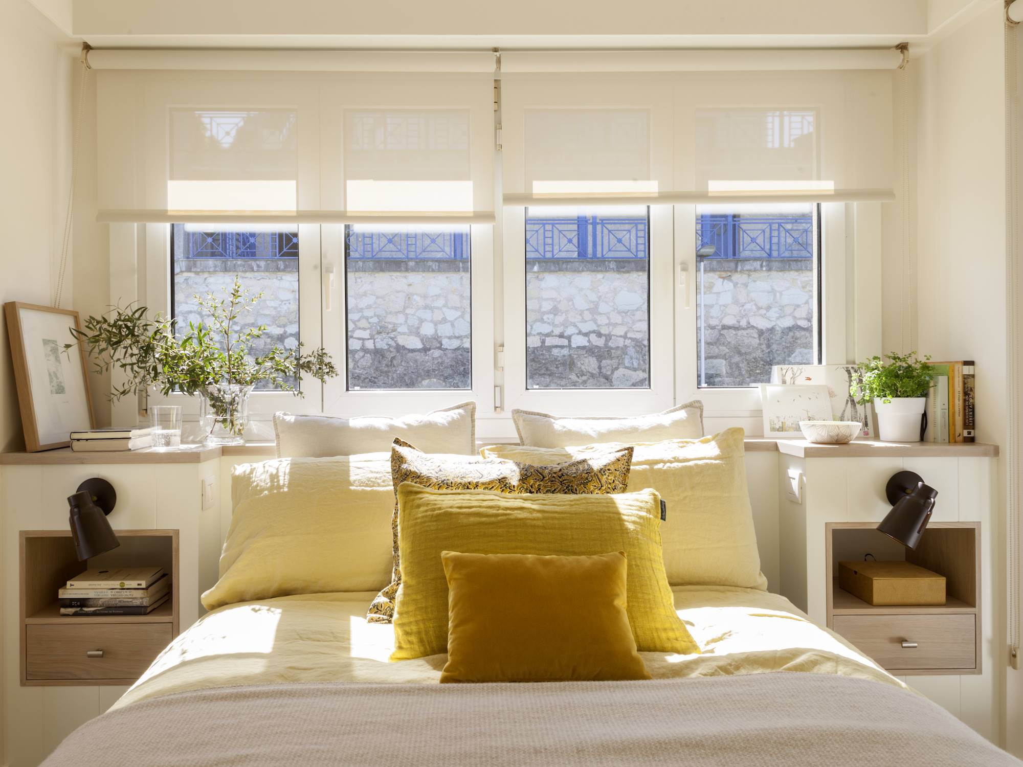 Dormitorio con cama con cojines ocre debajo la ventana.