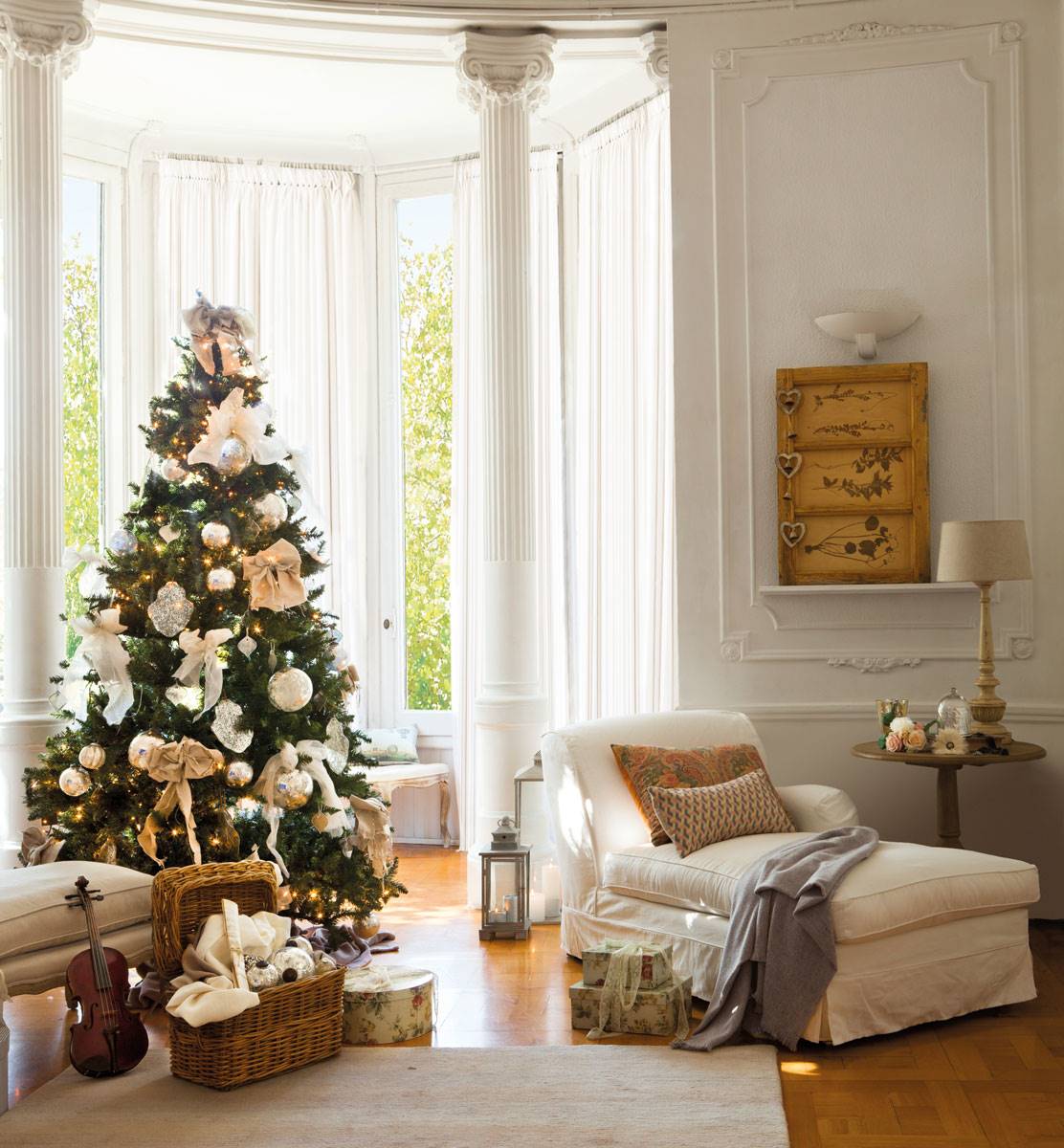 Salón elegante con molduras blancas, chaise longue, cesta y violín en el suelo y árbol de. Navidad decorado con bolas plateadas y lazos.