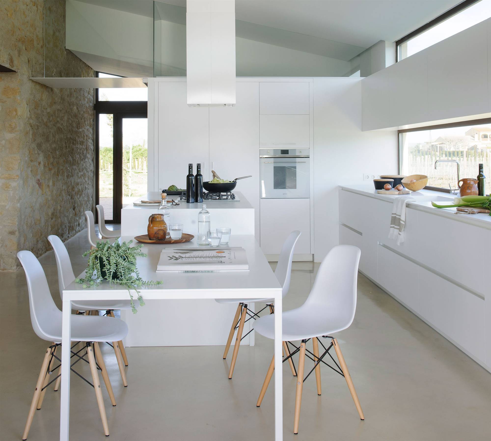 Cocina de diseño con mobiliario blanco, isla con campana extractora original y mesa que hace de office en el mismo color. 