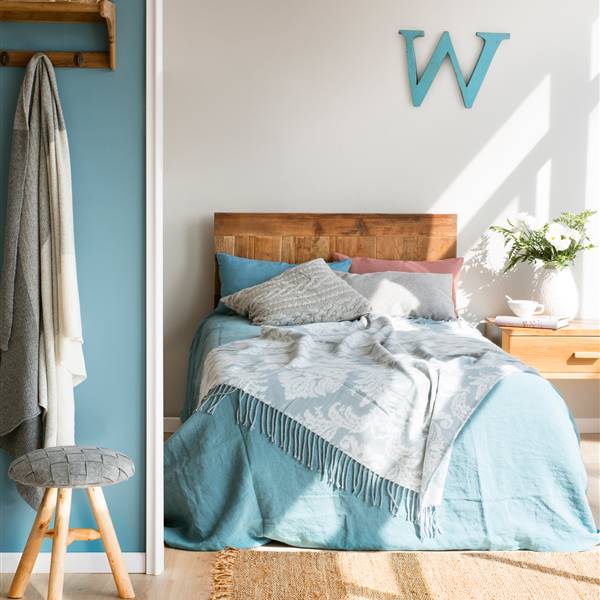 Dormitorio con pared gris piedra y azul, con cabecero y mesita de noche de madera, letra W decorativa, ropa de cama con estampado damasco, taburete y colgador