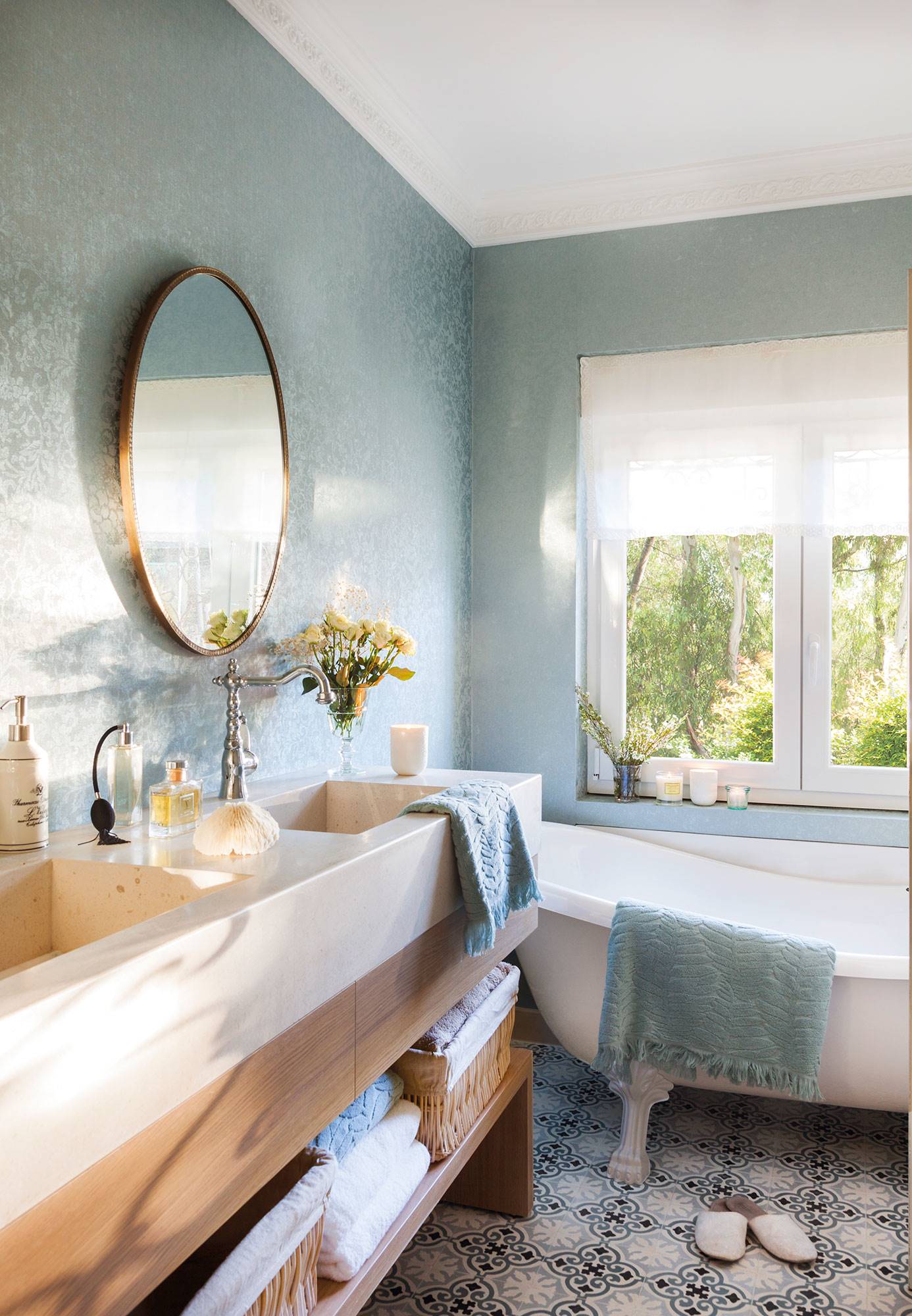 Baño con papel pintado efecto textil de color azul, mueble bajolavabo de madera y bañera con patas. 