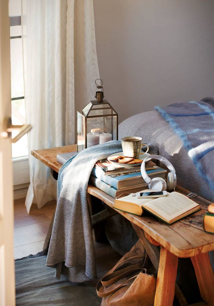 Dormitorio con banqueta de madera con libros y fanalillos sobre ella. 