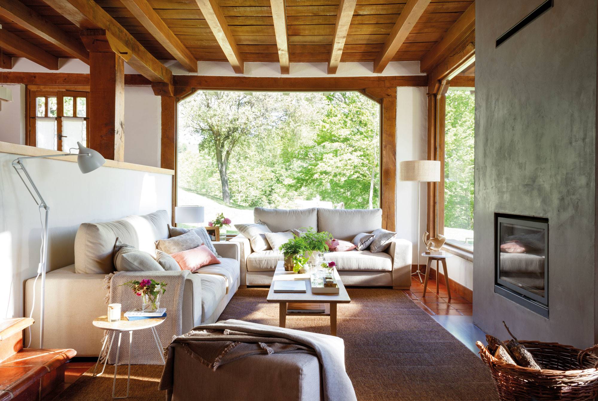 Salón rústico con techos y vigas de madera, sofá esquinero en gris y chimenea. 