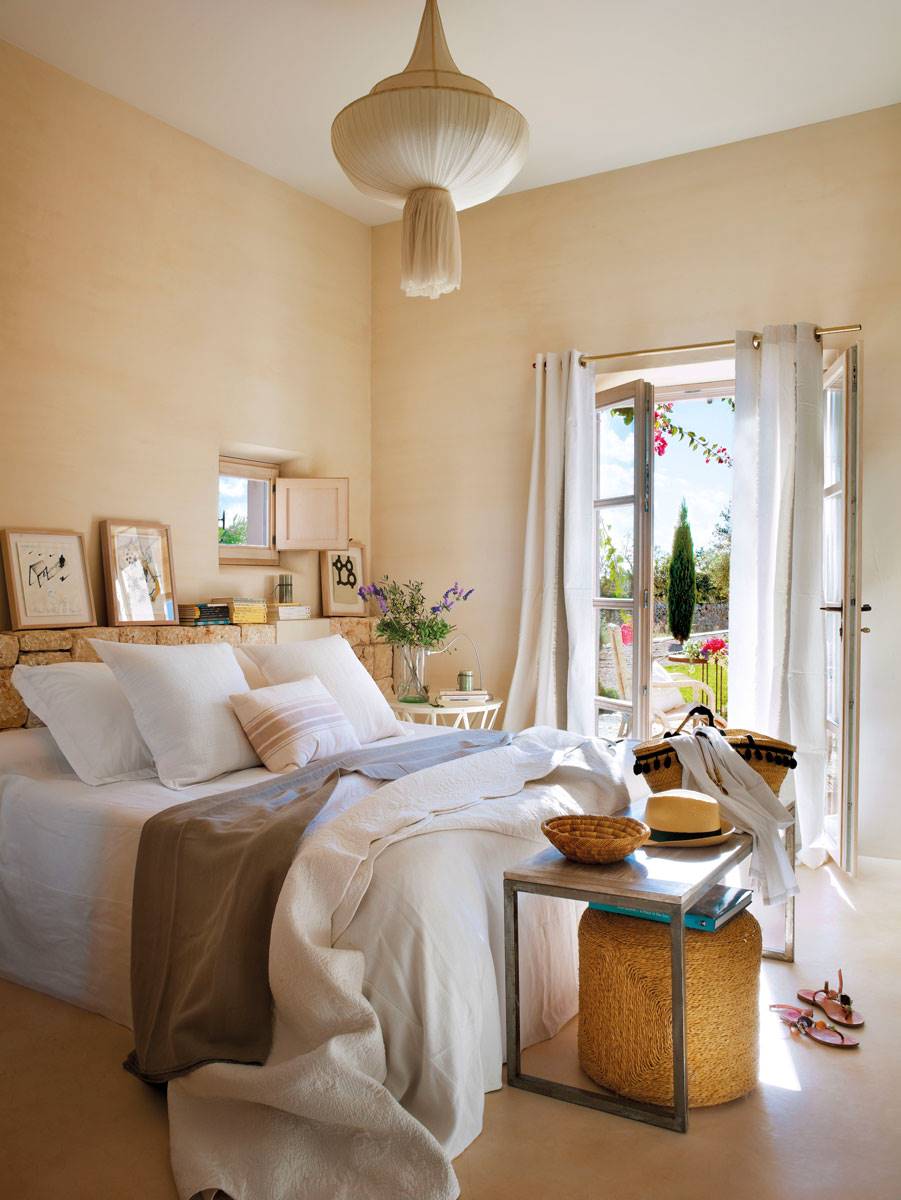 Dormitorio en tonos ocres con cabecero de piedra y lámpara de techo con pavimento de cemento pulido.
