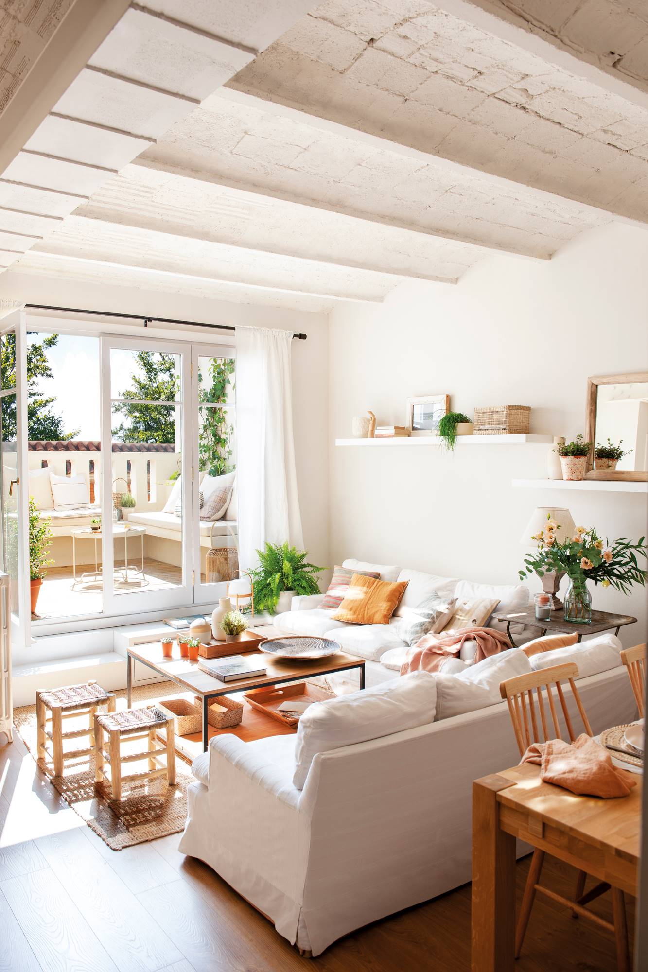Salón pequeño con mesa de centro de madera y hierro, dos sofás blancos y ventana con acceso a la terraza. 