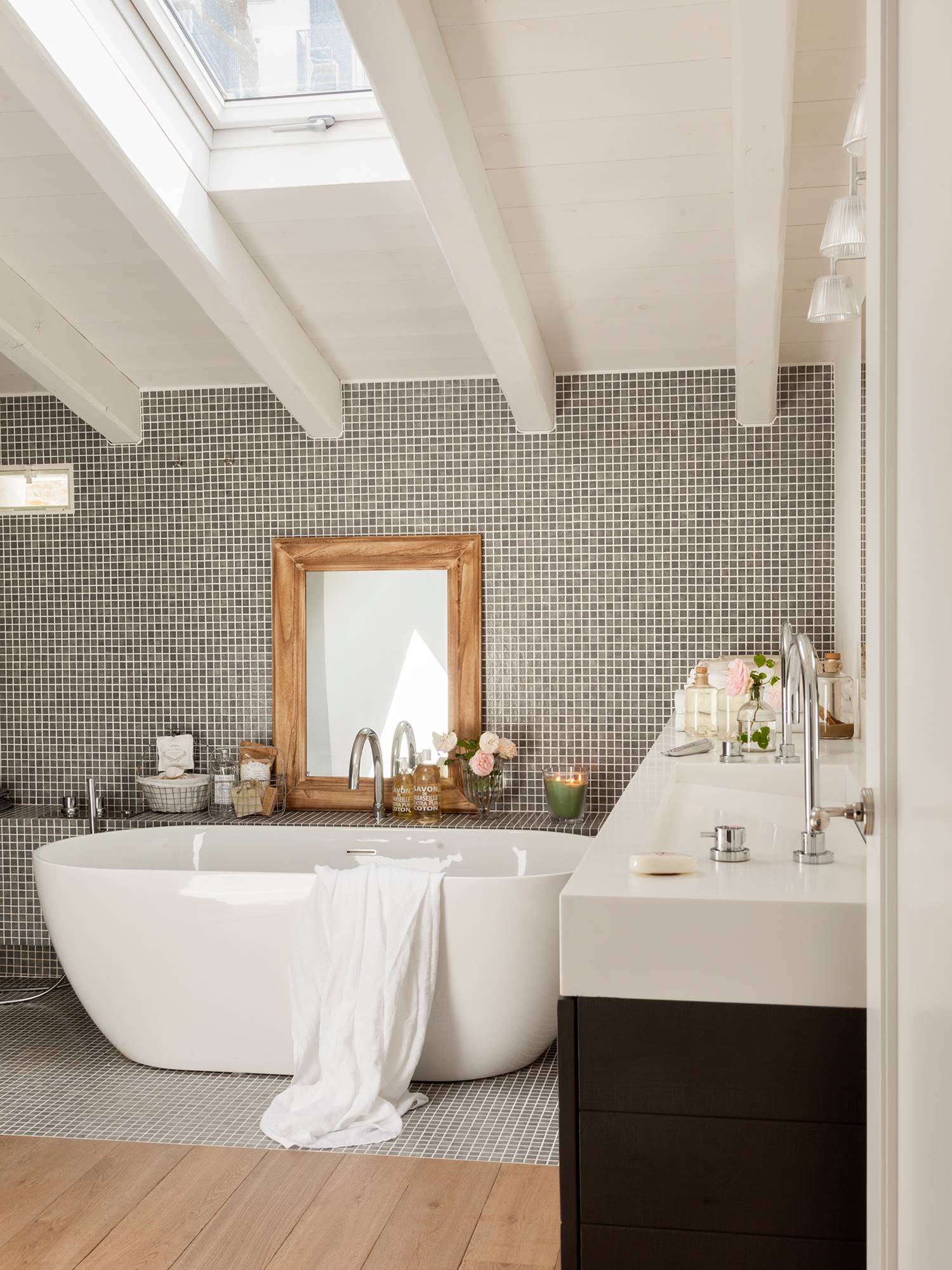 Baño reformado con bañera extenta, espejo de madera encima, azulejos pequeños y gresite. 