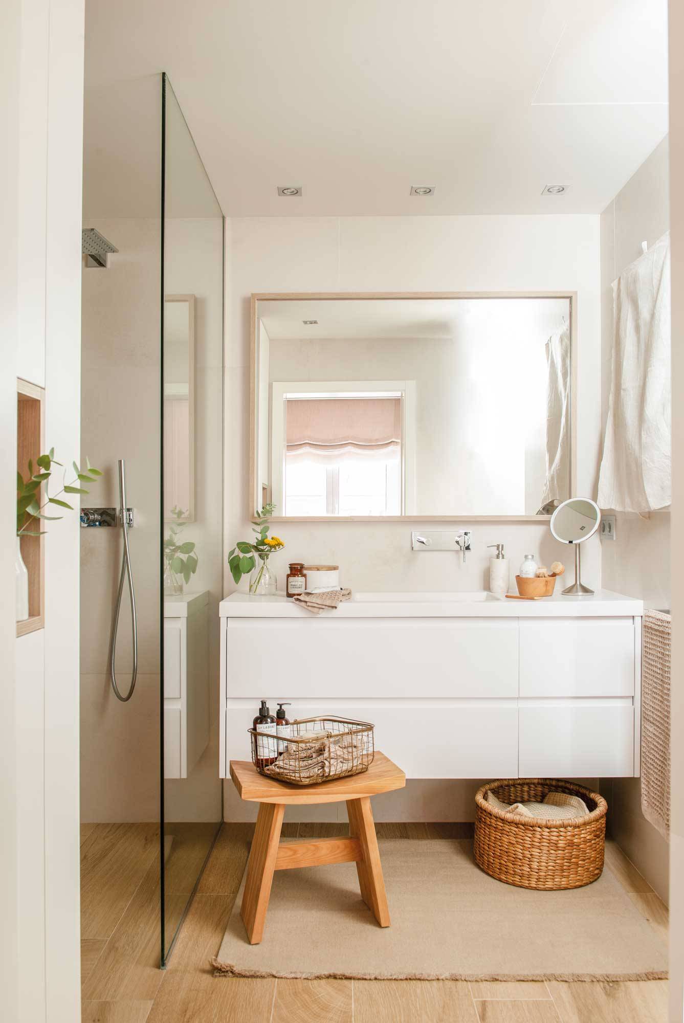 Baño reformado con suelo de madera, ducha con mampara, alfombra y mueble bajolavabo blanco.