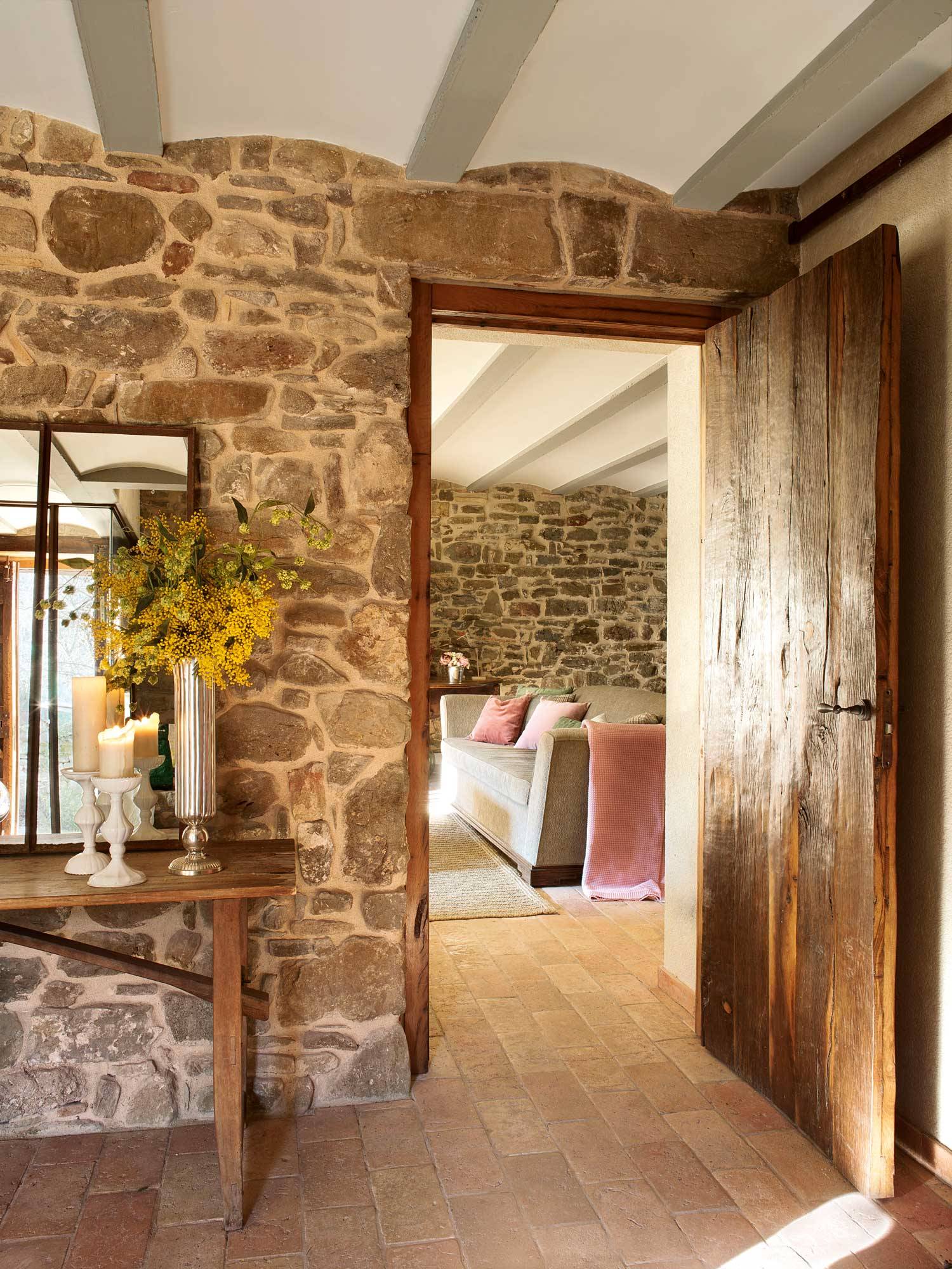 Recibidor rústico con pared de piedra, puerta de madera, consola de madera, espejo, velas y mimosas.
