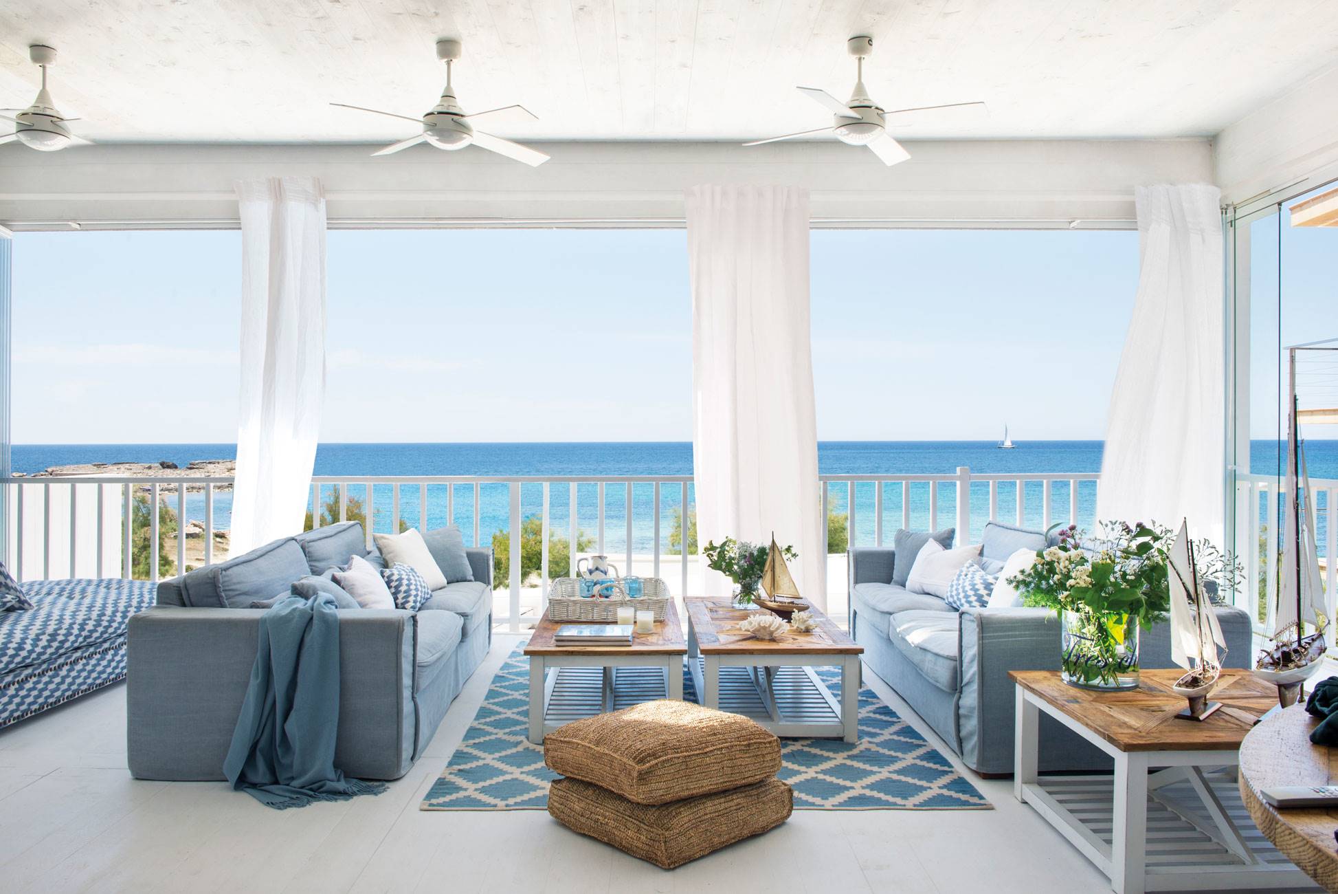 Salón de verano con vistas al mar con sofa´s azules y ventiladores de techo. 