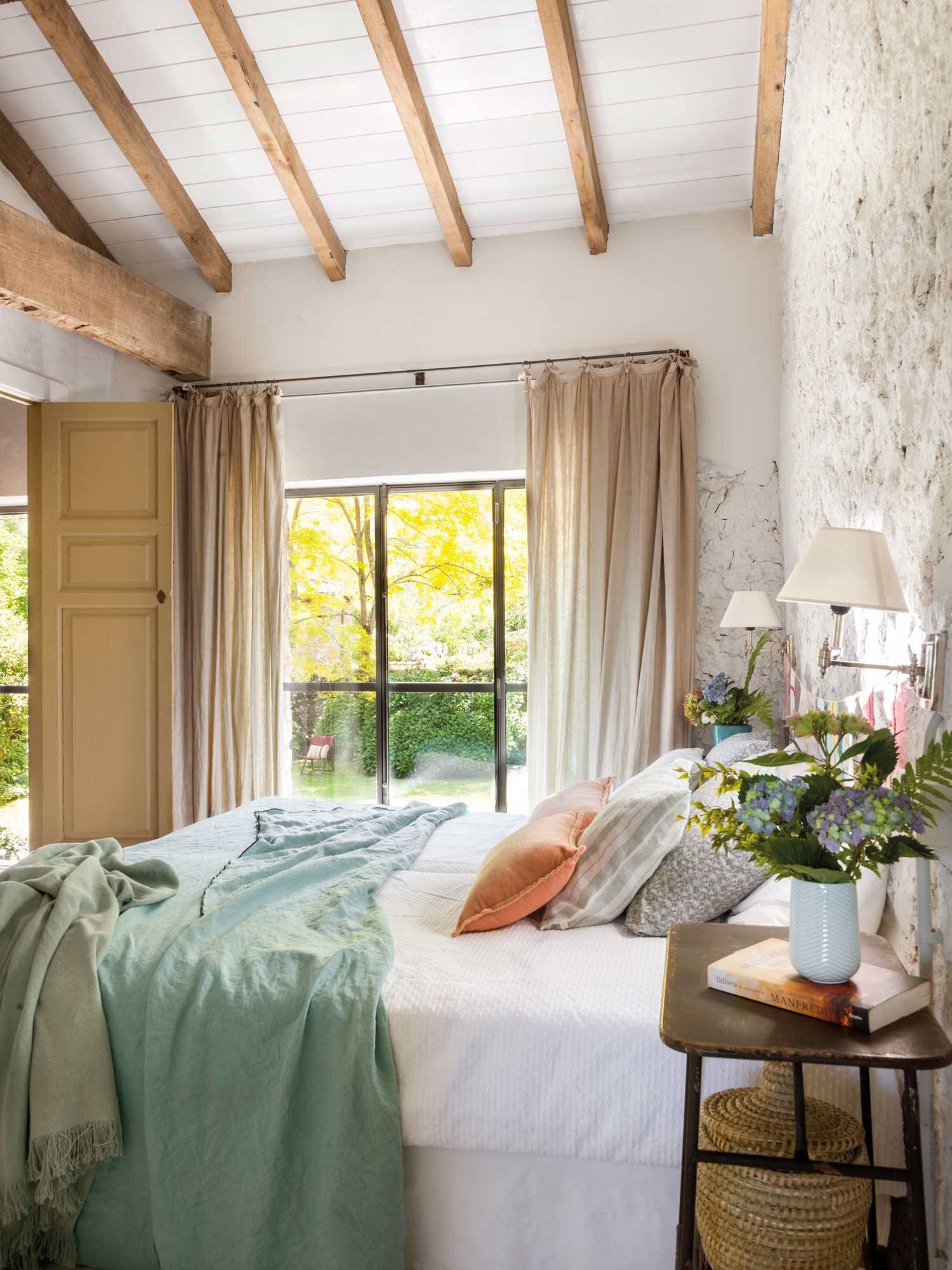 Dormitorio de verano con paredes de piedra blancas y ropa de cama en blanco y tonos pastel. 
