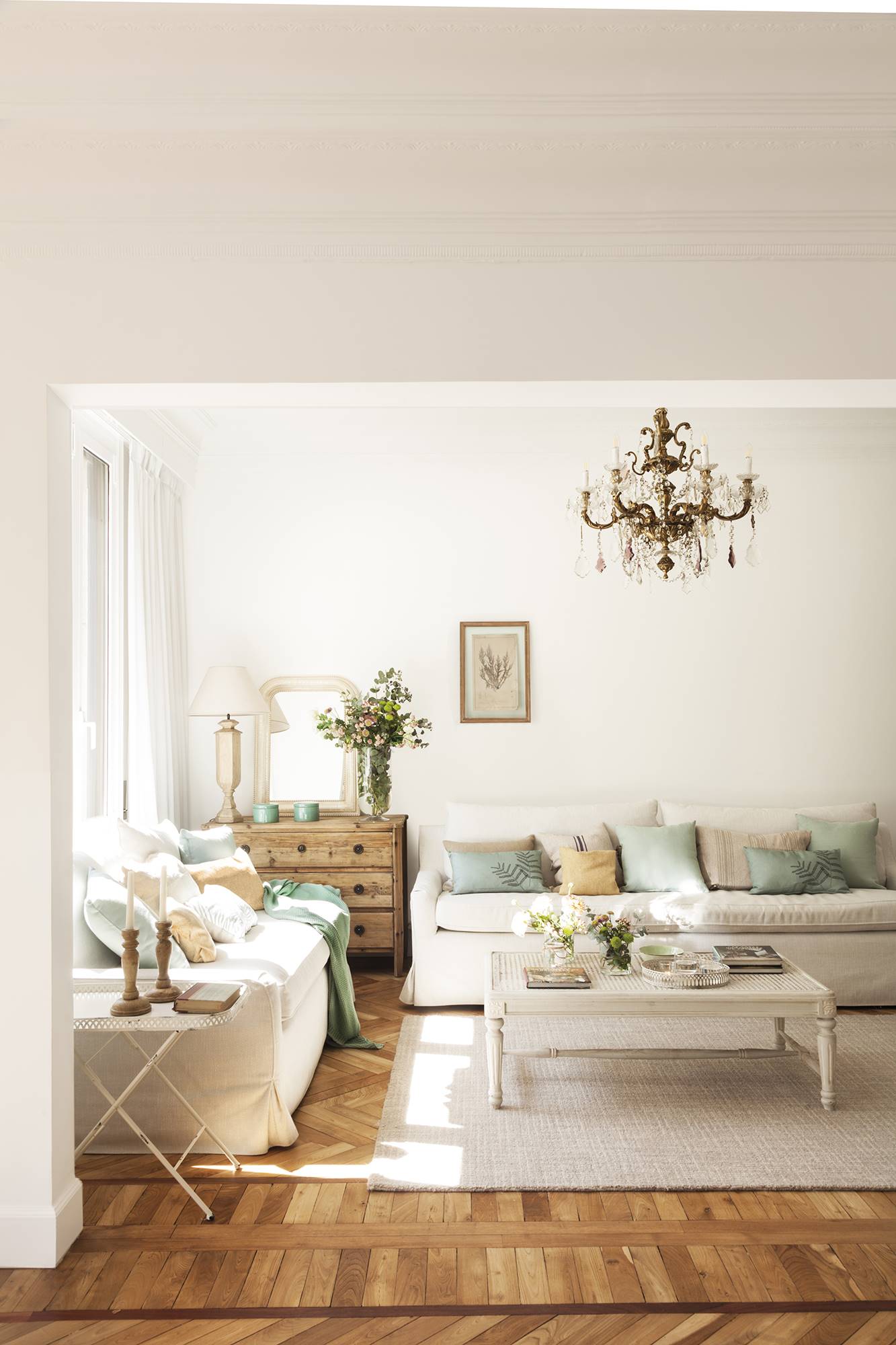 Salón blanco con dos sofás, mesa de centro clásica y lámpara de araña.