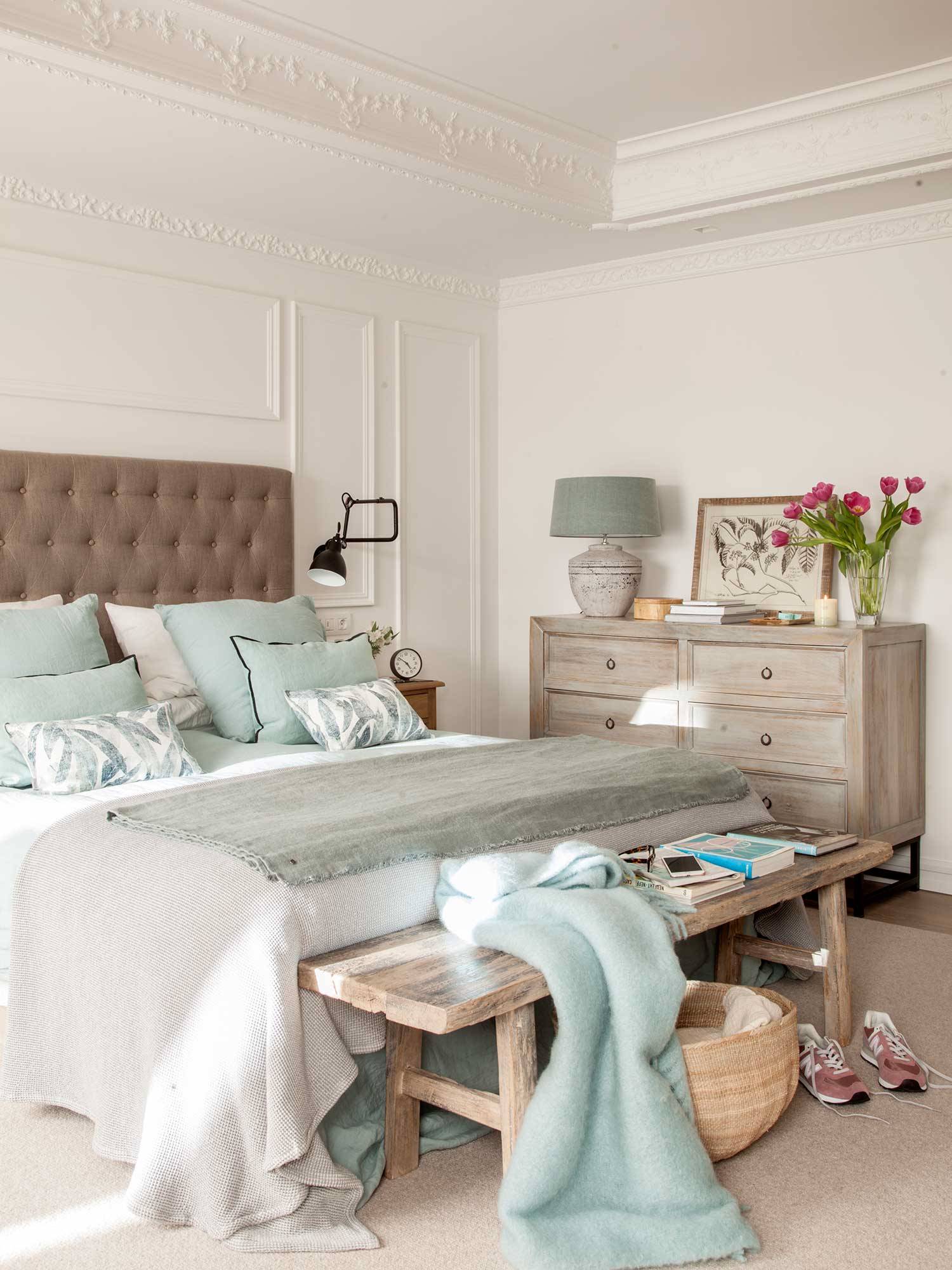 Dormitorio con molduras, cabecero capitoné marrón, cómoda y banco de madera.