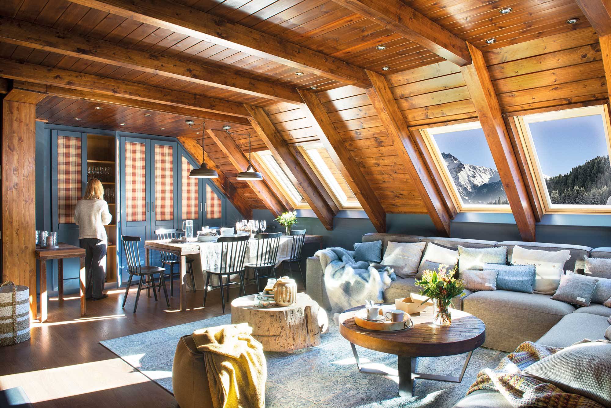 Salón de casa de montaña con paredes y techos de madera, gran sofá esquinero gris y armario azul con tela de cuadros.