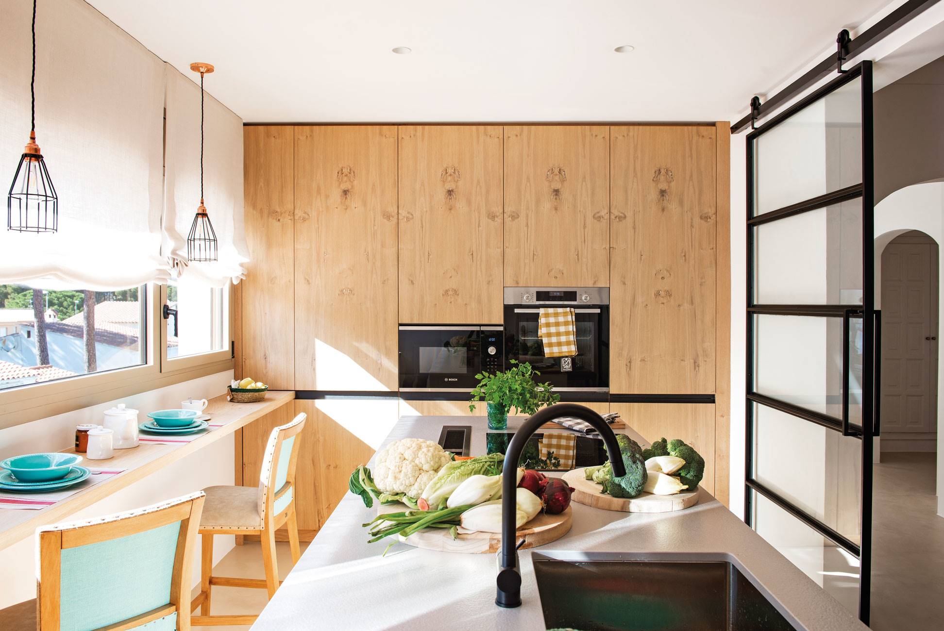 Moderna cocina blanca y de madera con balda en la pared a modo de mesa para desayunar y puerta de cristal ocrredera.