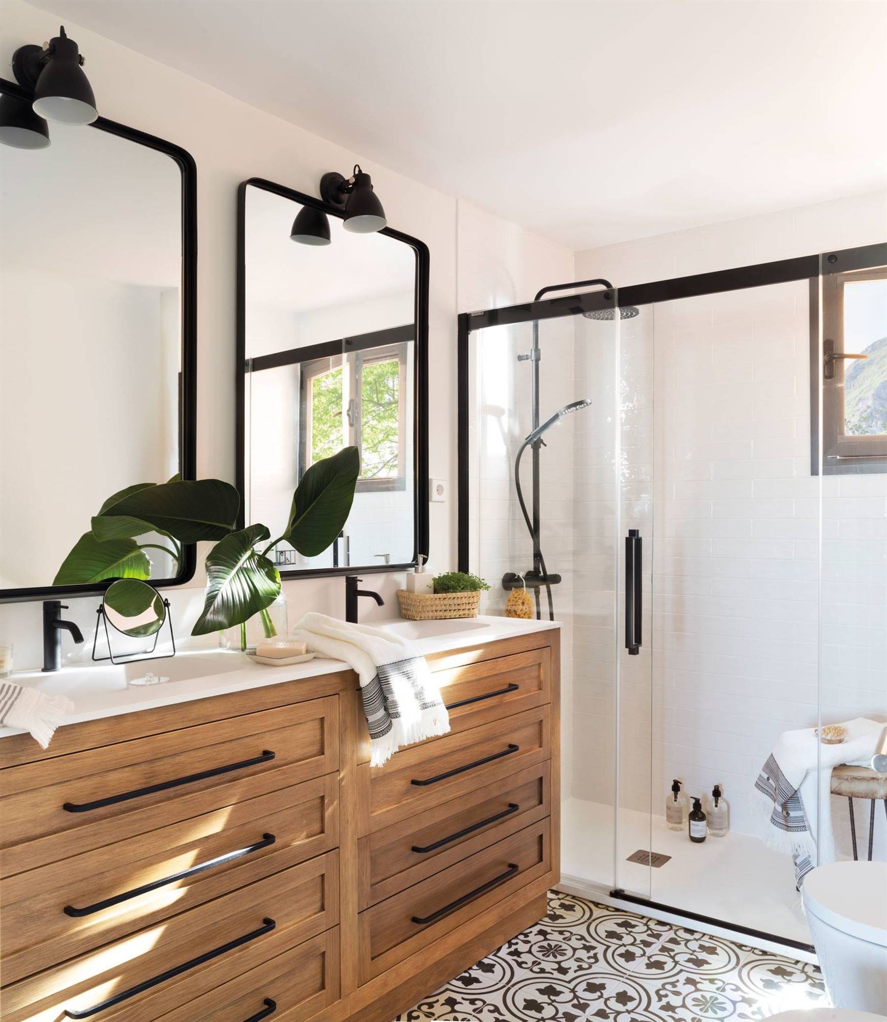 Baño con ducha, dos lavabos empotrados en un mueble de madera, suelo de baldosa hidráulica y espejos con marco metálico negro a juego con los grifos.