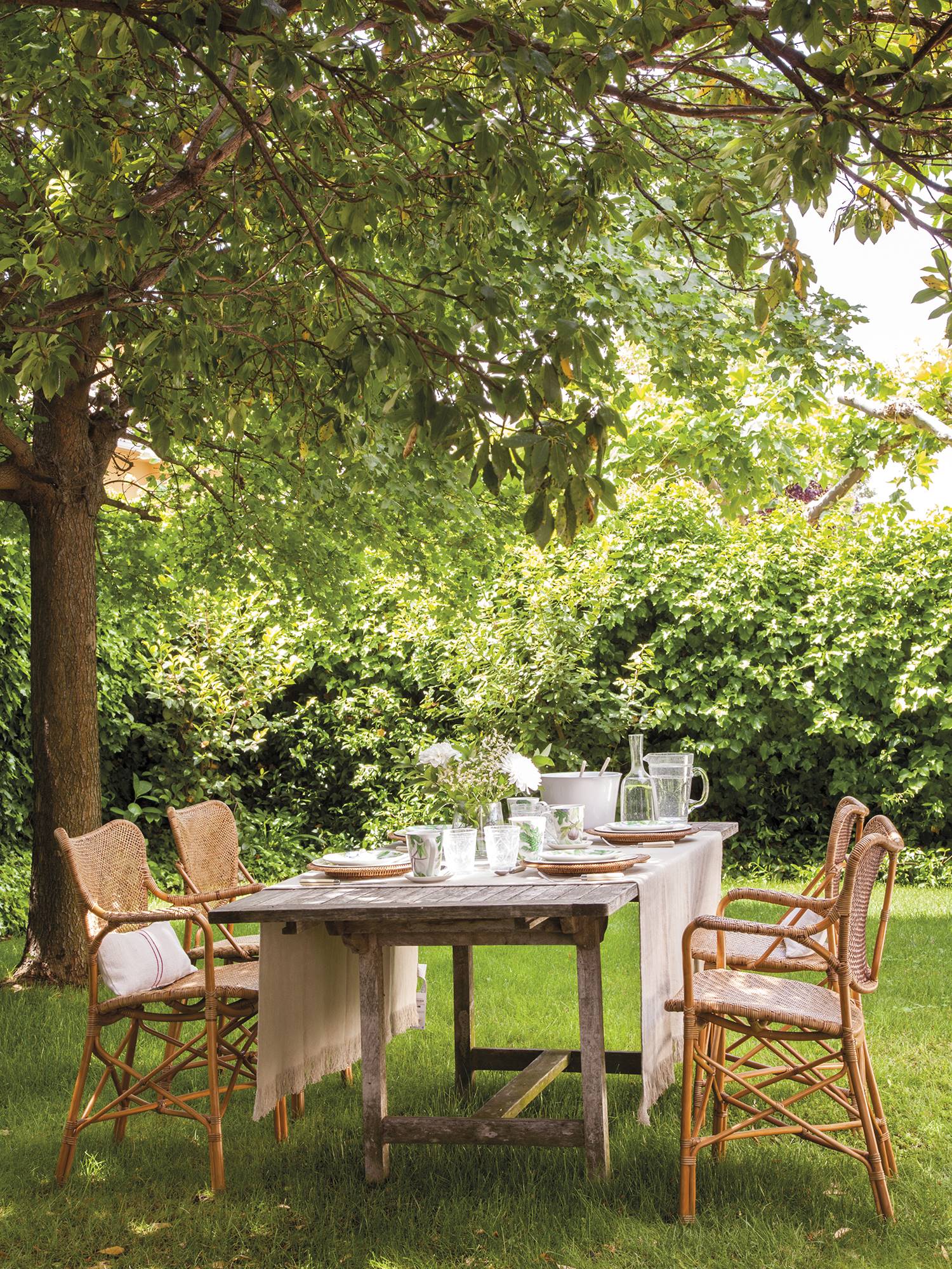 Comedor exterior bajo los árboles con sillas de ratán y mantel en tonos beige. 