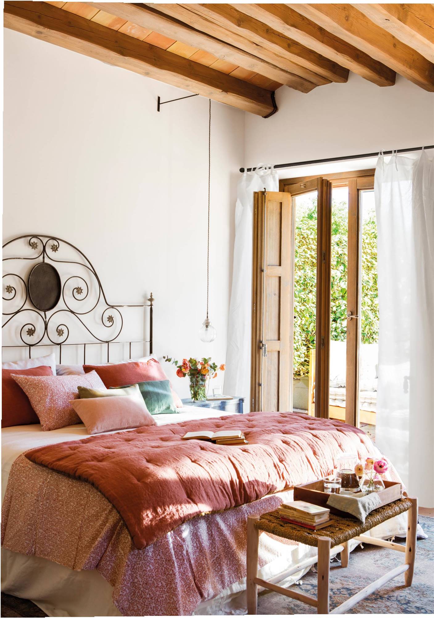 Dormitorio con cabecero de forja, ropa de cama rosa y pie de madera y fibras naturales al pie de la cama. 