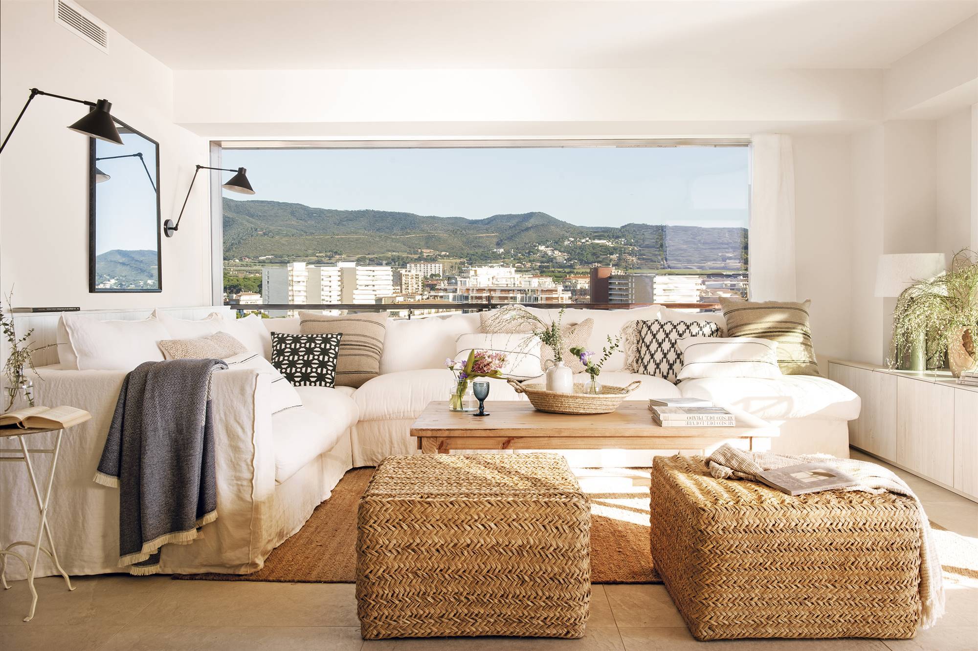 Salón con sofá esquinero de lino blanco, mesa de centro de madera, dos pufs de fibras, alfombra de fibras y terraza con vistas panorámicas al mar