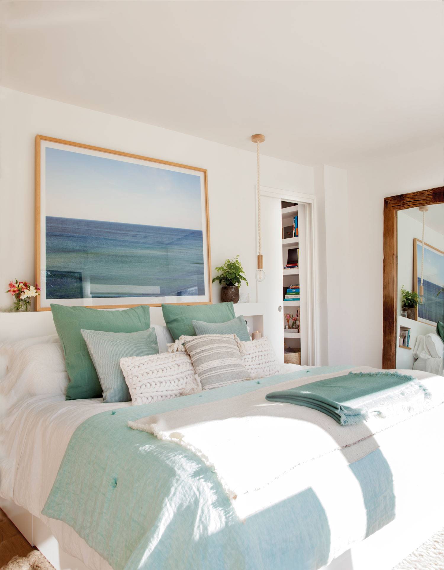 Dormitorio con cabecero de obra blanco y hornacinas, un cuadro grande de un paisaje marino y lámparas suspendidas.