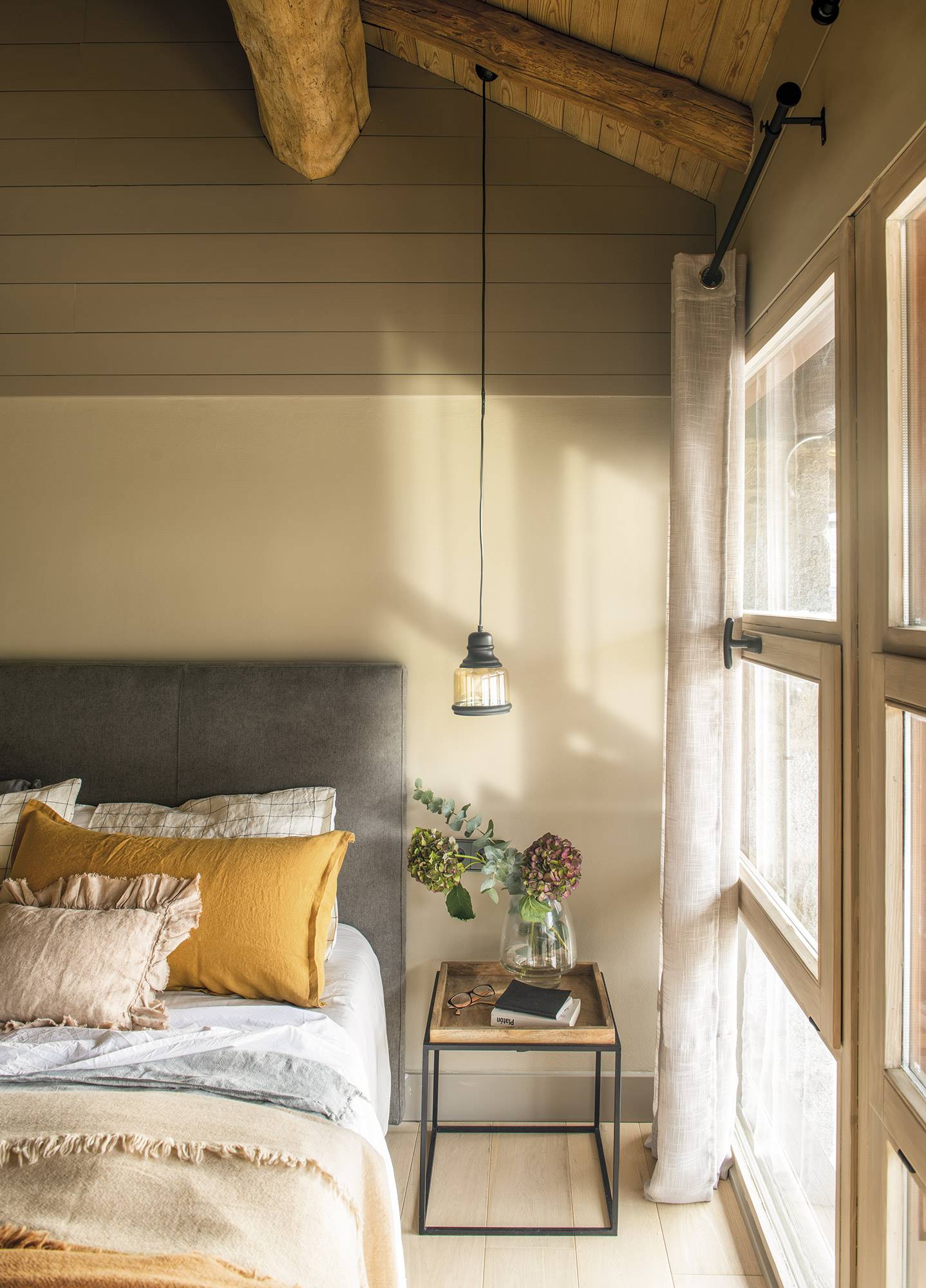 Dormitorio de estilo rústico con vigas de madera, cabecero tapizado de algodón gris y mesita de noche de madera.