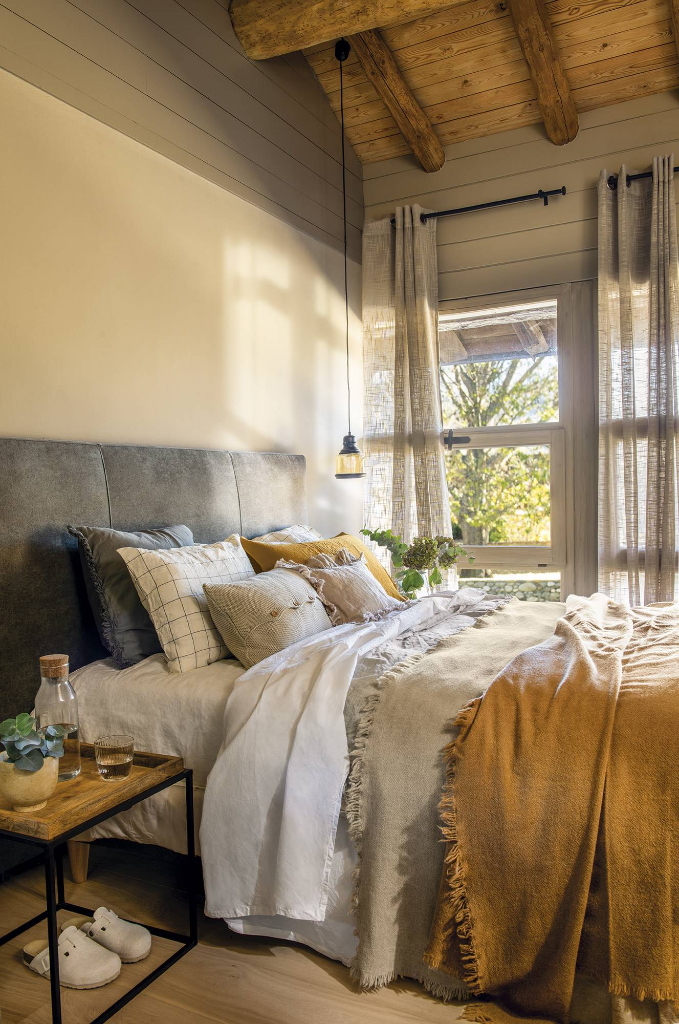 Dormitorio rústico con cabecero tapizado de gris, vigas de madera en el techo y ropa de cama gris y mostaza. 