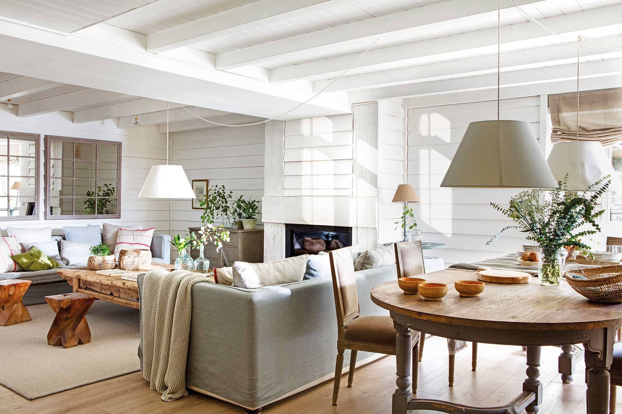Salón de casa de montaña de color blanco con mesa de comedor de madera y sofás en gris.