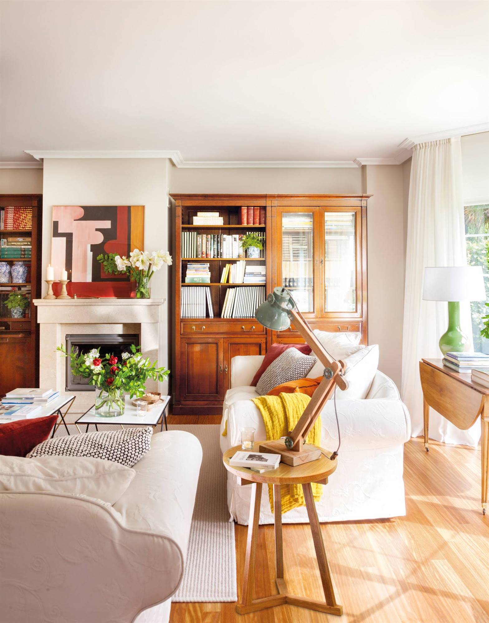 Salón con librería tipo vitrina de madera, dos sofás blancos y chimenea con cuadro encima. 