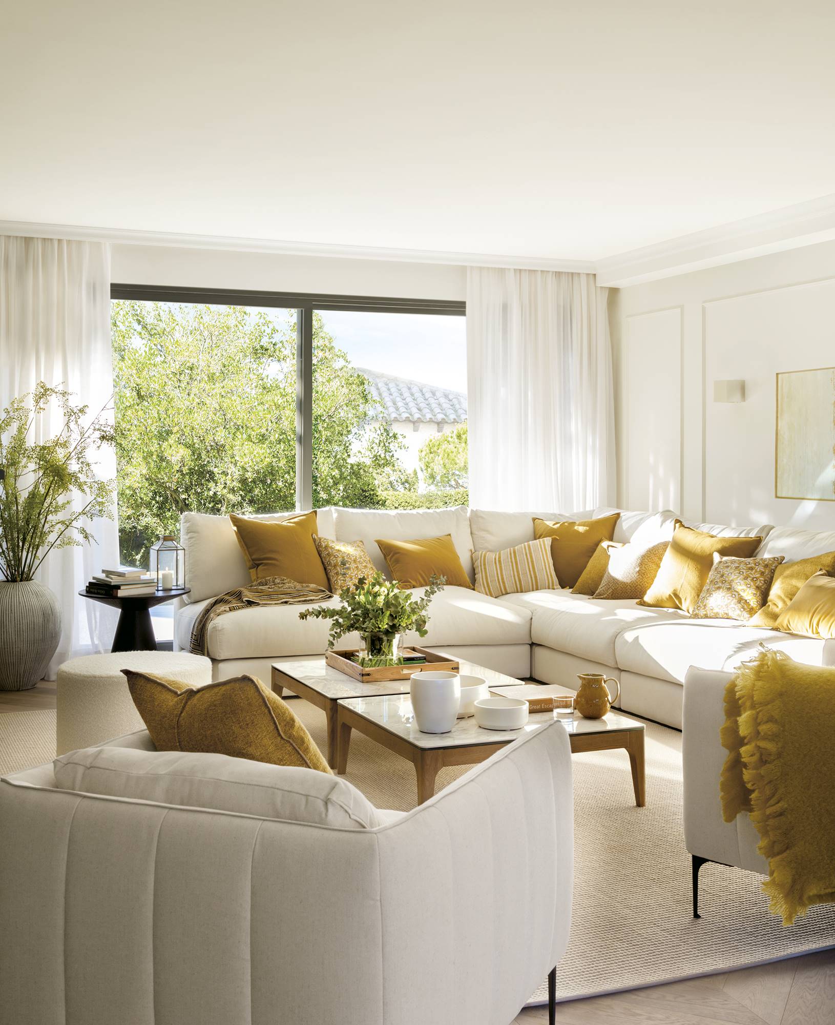 Zona de estar con sofá esquinero blanco, paredes blancas con molduras y cortinas de lino.