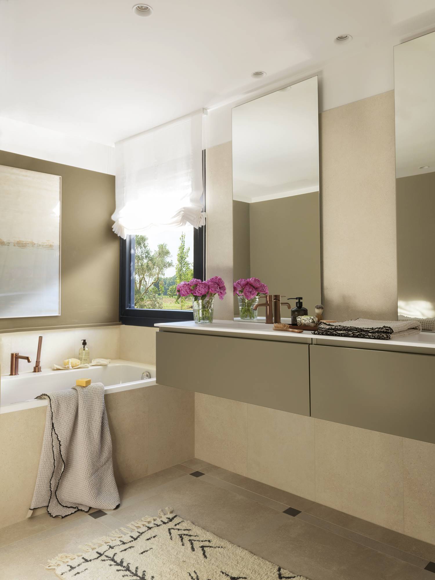 Baño en suite con bañera y mueble bajolavabo volado, estor blanco, grifería en acabado cobre y espejos sin marco.