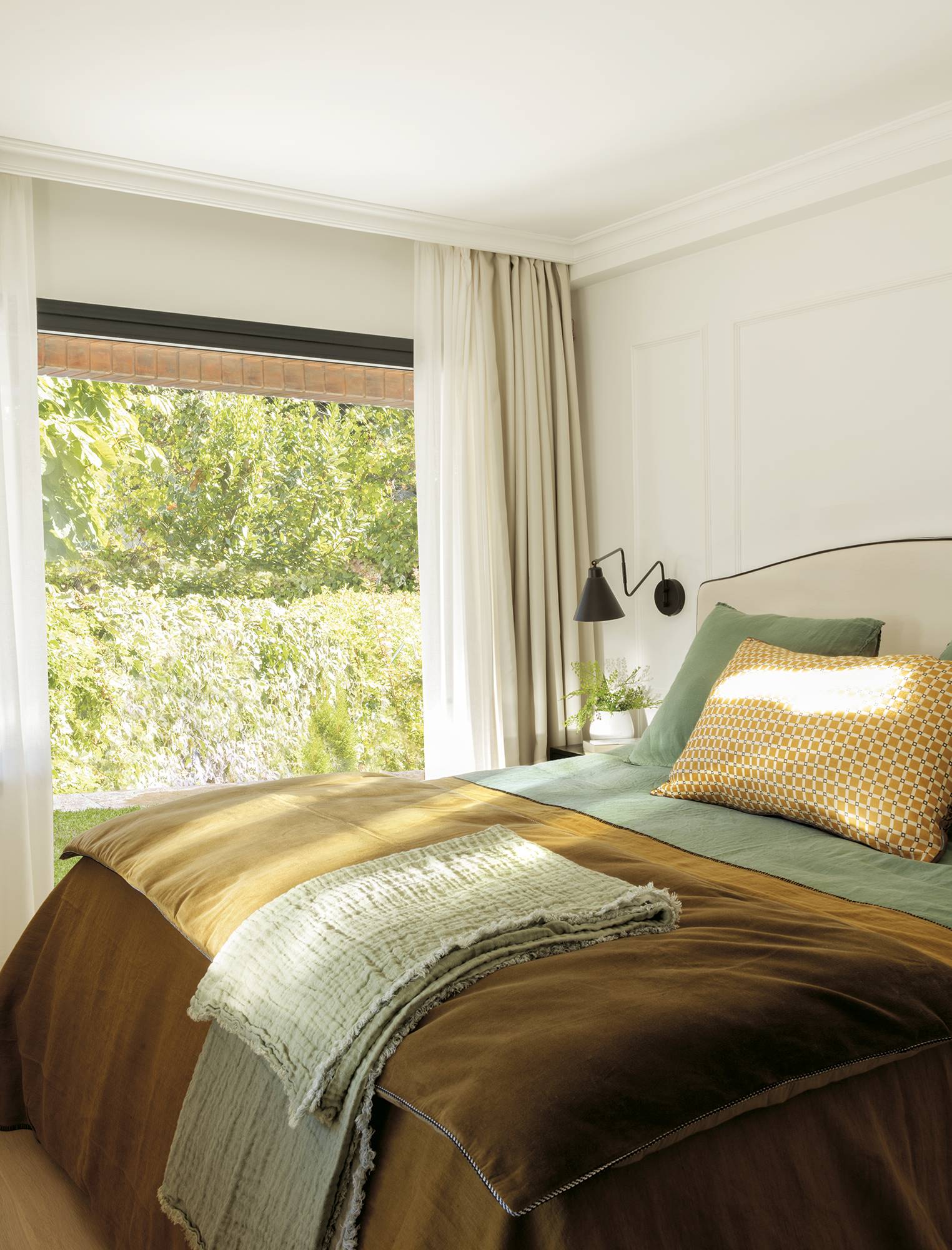 Dormitorio cálido y elegante con cabecero tapizado, cortinas blancas y ropa de cama verde y amarillo mostaza.