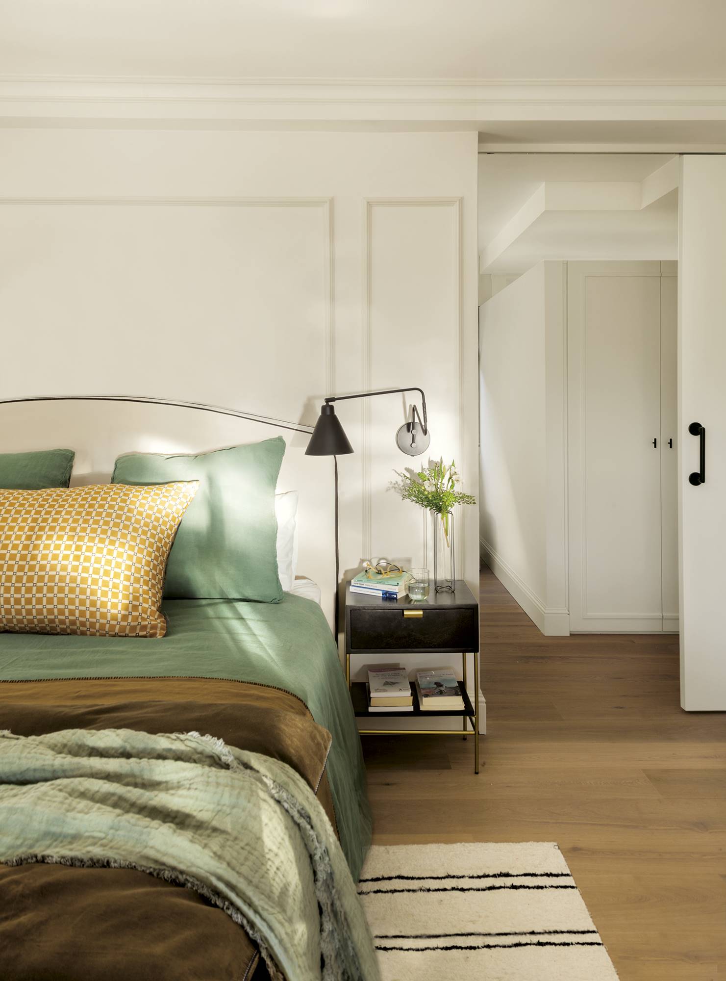 Dormitorio con vestidor, decorado en blanco con molduras, cabecero tapizado, ropa de cama verde y apliques de lectura negros.