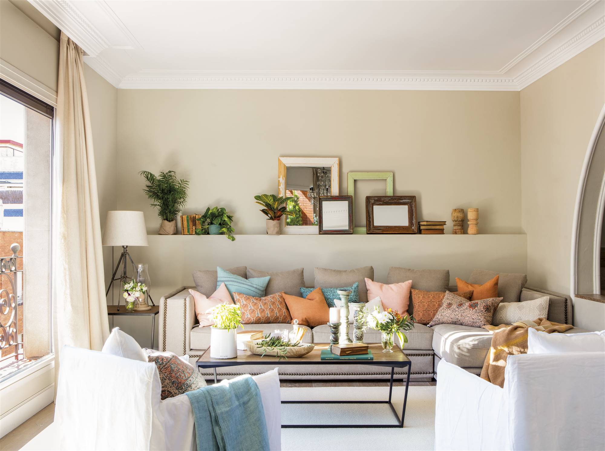 Salón con sofá lleno de cojines de colores con chaise longue, butacas blancas y muerte tras el sofá con cuadros. 