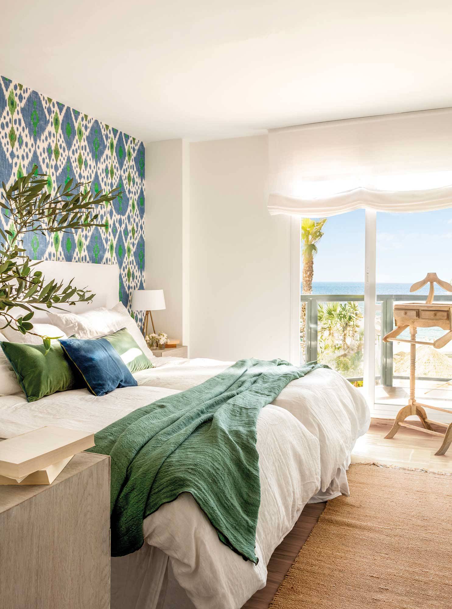 Dormitorio de verano con papel pintado azul y verde en el cabecero y ropa de cama blanca. 