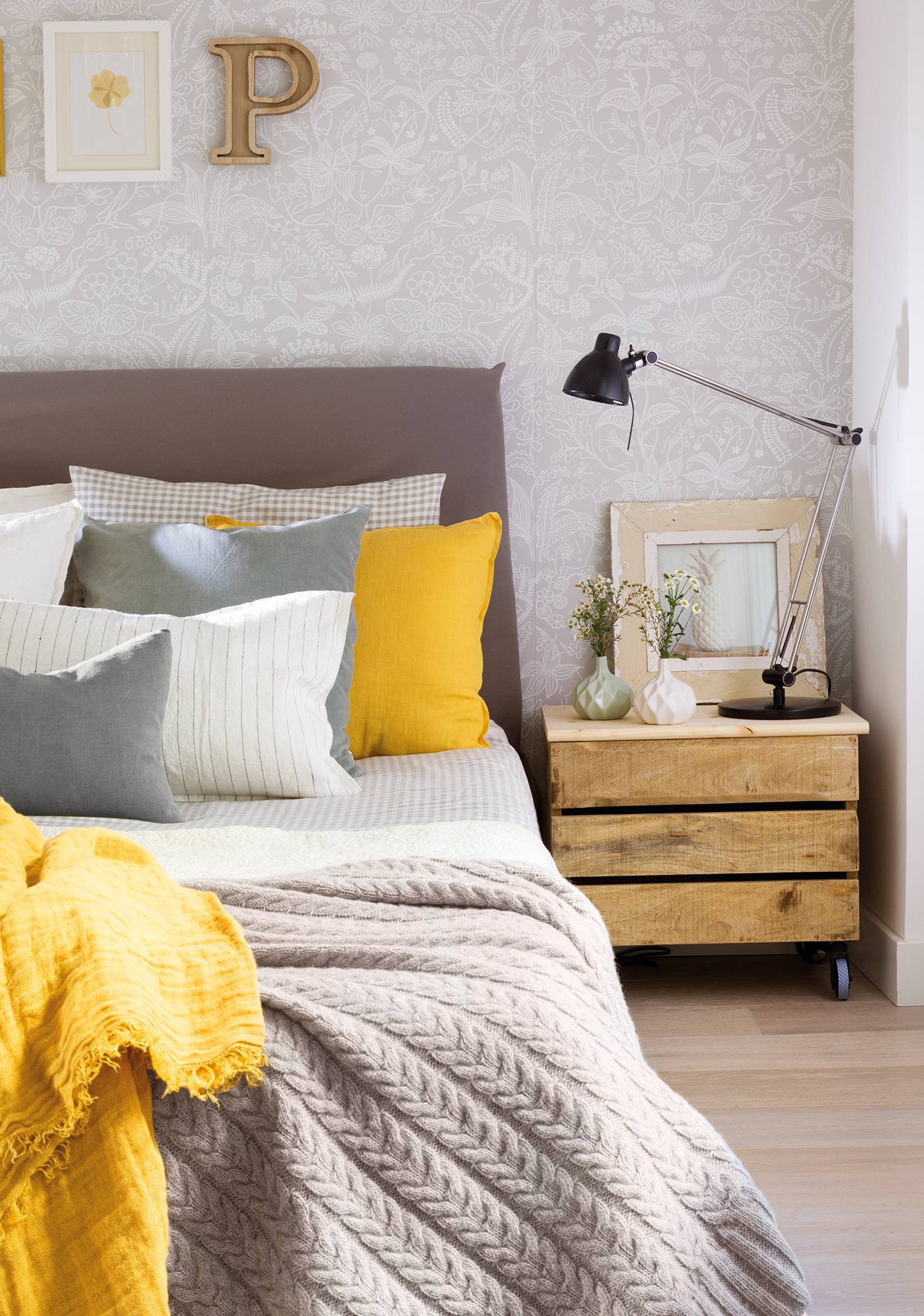 Semitoma de dormitorio con papel pintado y cama con cojines y mesilla de noche hecha con caja de madera.