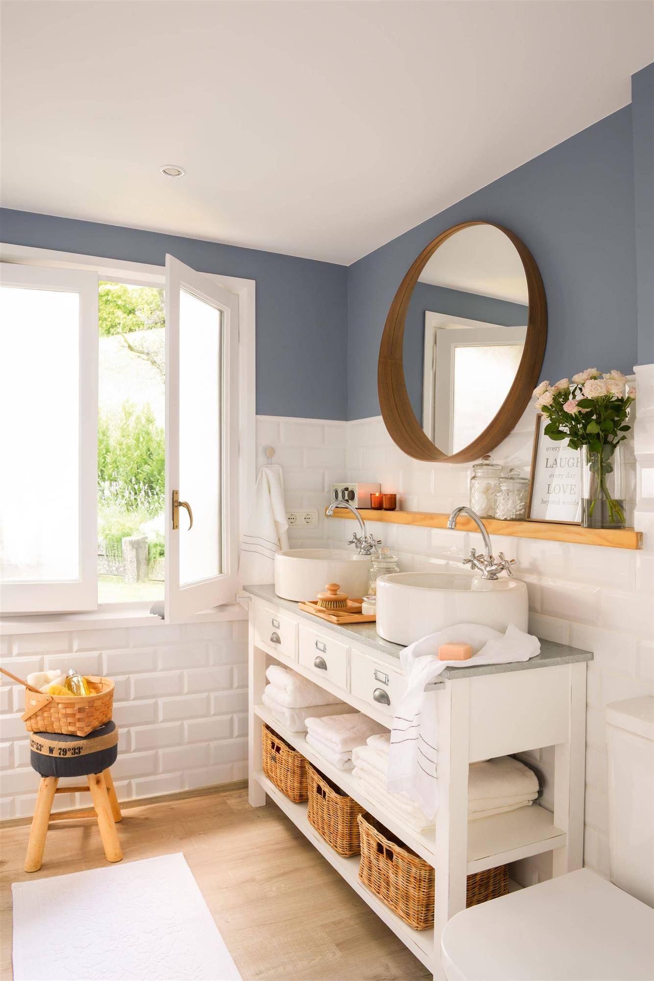 Baño con azulejos blancos a media altura y pintura azul. Espejo redondo y mueble bajolabavo de madera blanco. 