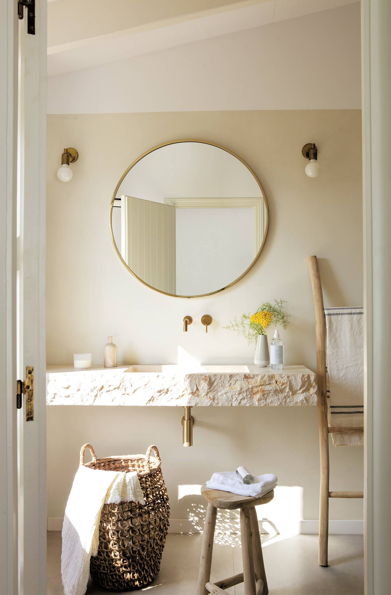 Baño con lavabo de piedra, grifería dorada, espejo redondo y escalera de madera