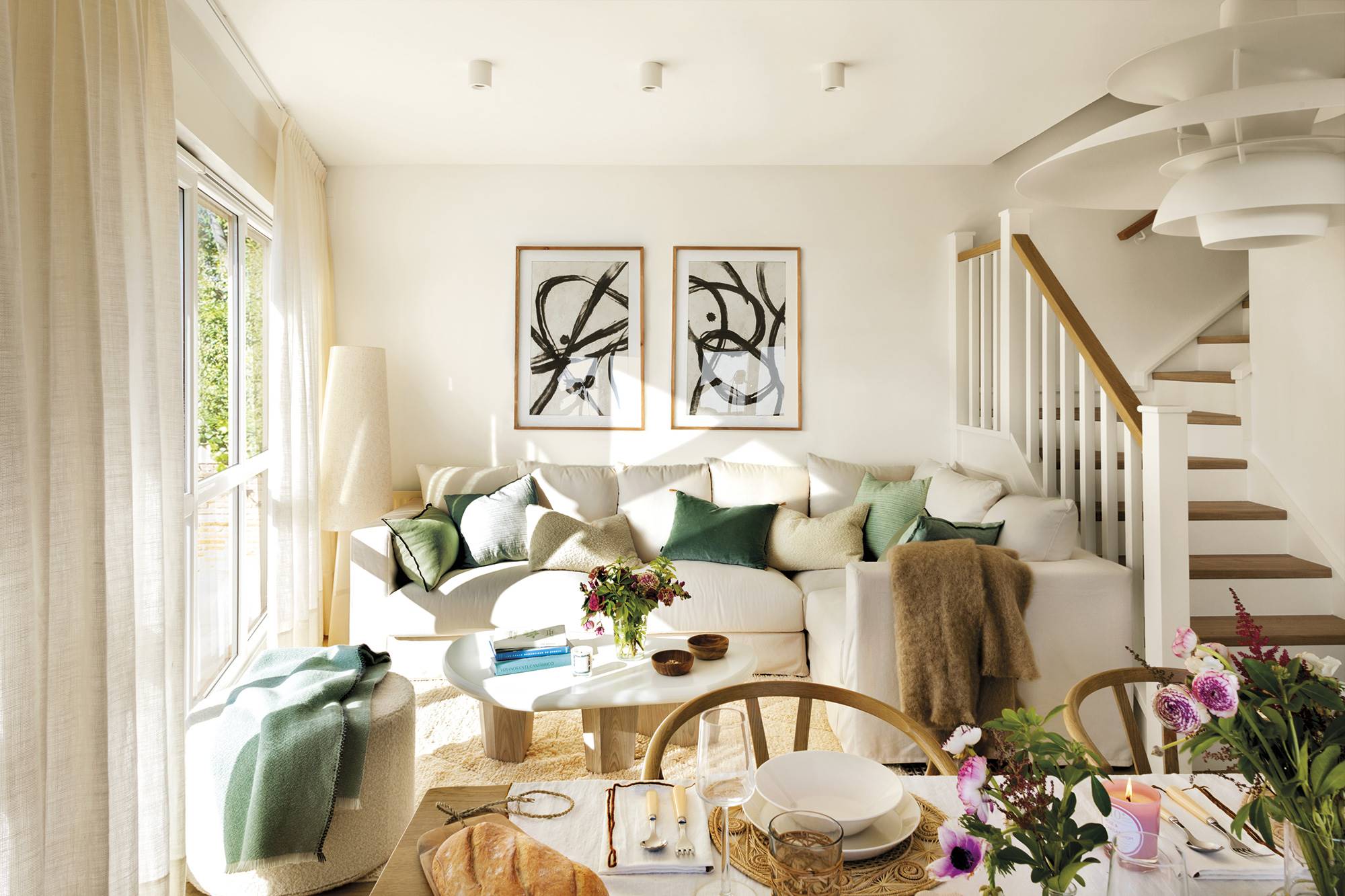 Salón con sofá blanco, sofás verdes, cuadros abstractos y escalera.